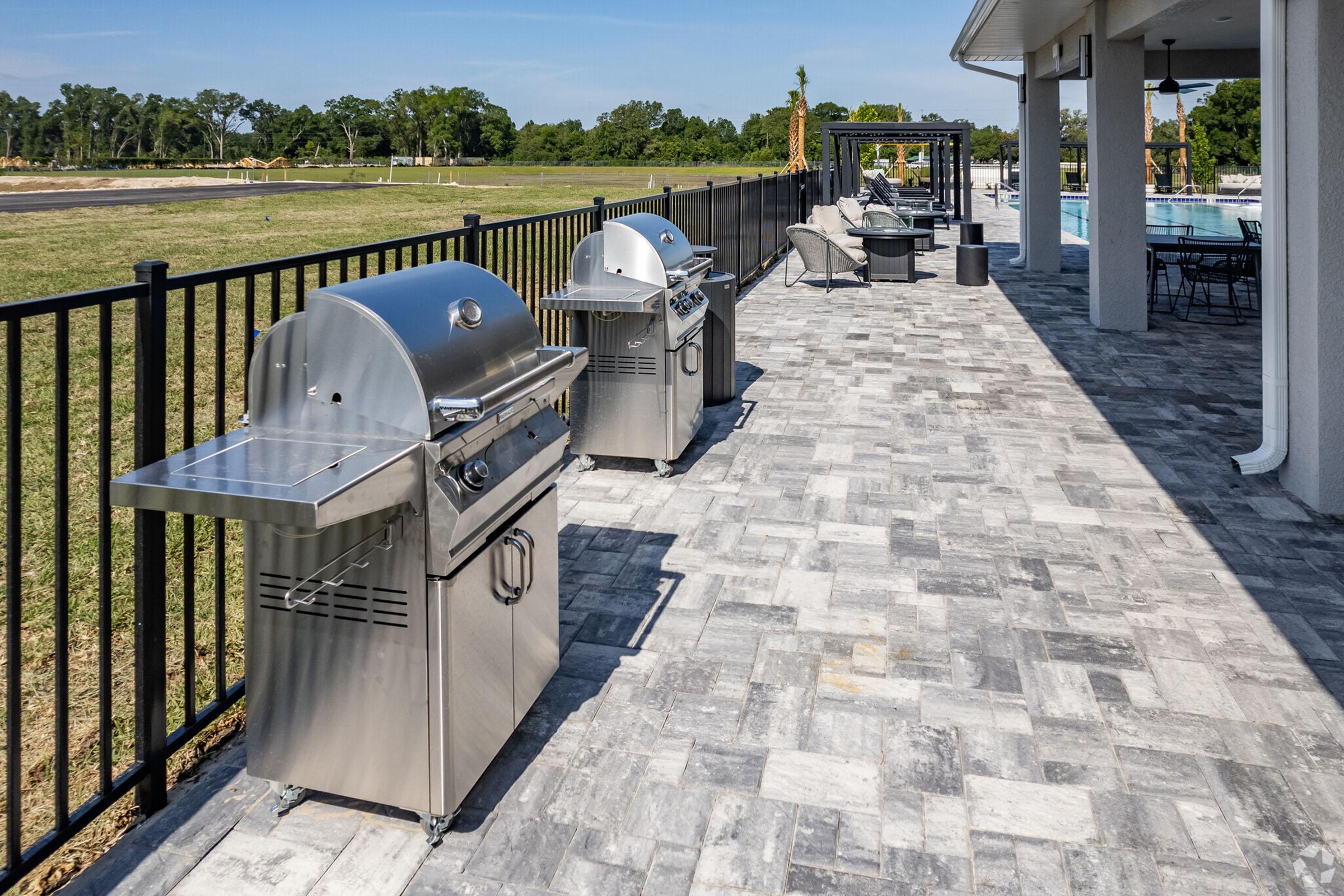 A patio area featuring two stainless steel grills on a paved surface. In the background, there is an open field with greenery, and a shaded seating area is visible. The sky is clear with a few clouds, creating a sunny atmosphere. The setting is ideal for outdoor cooking and social gatherings.