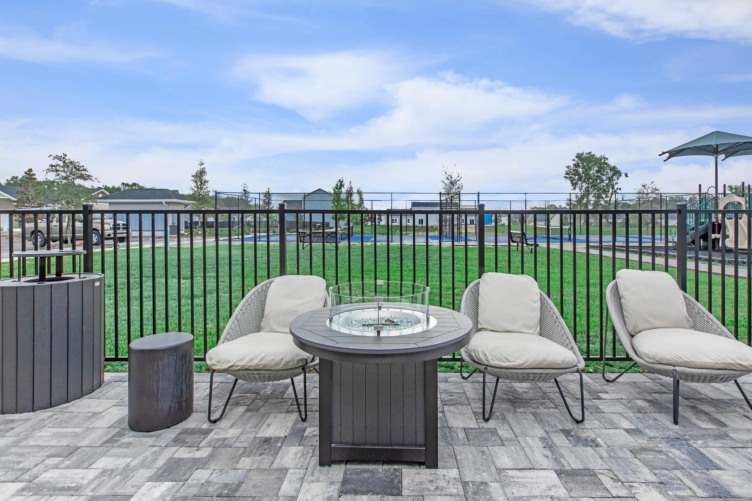 Outdoor seating area featuring three modern lounge chairs around a fire pit table. The setting includes a grassy lawn in the background with a fence, and a parasol nearby, creating a relaxing space ideal for gatherings or leisure. The sky is partly cloudy.