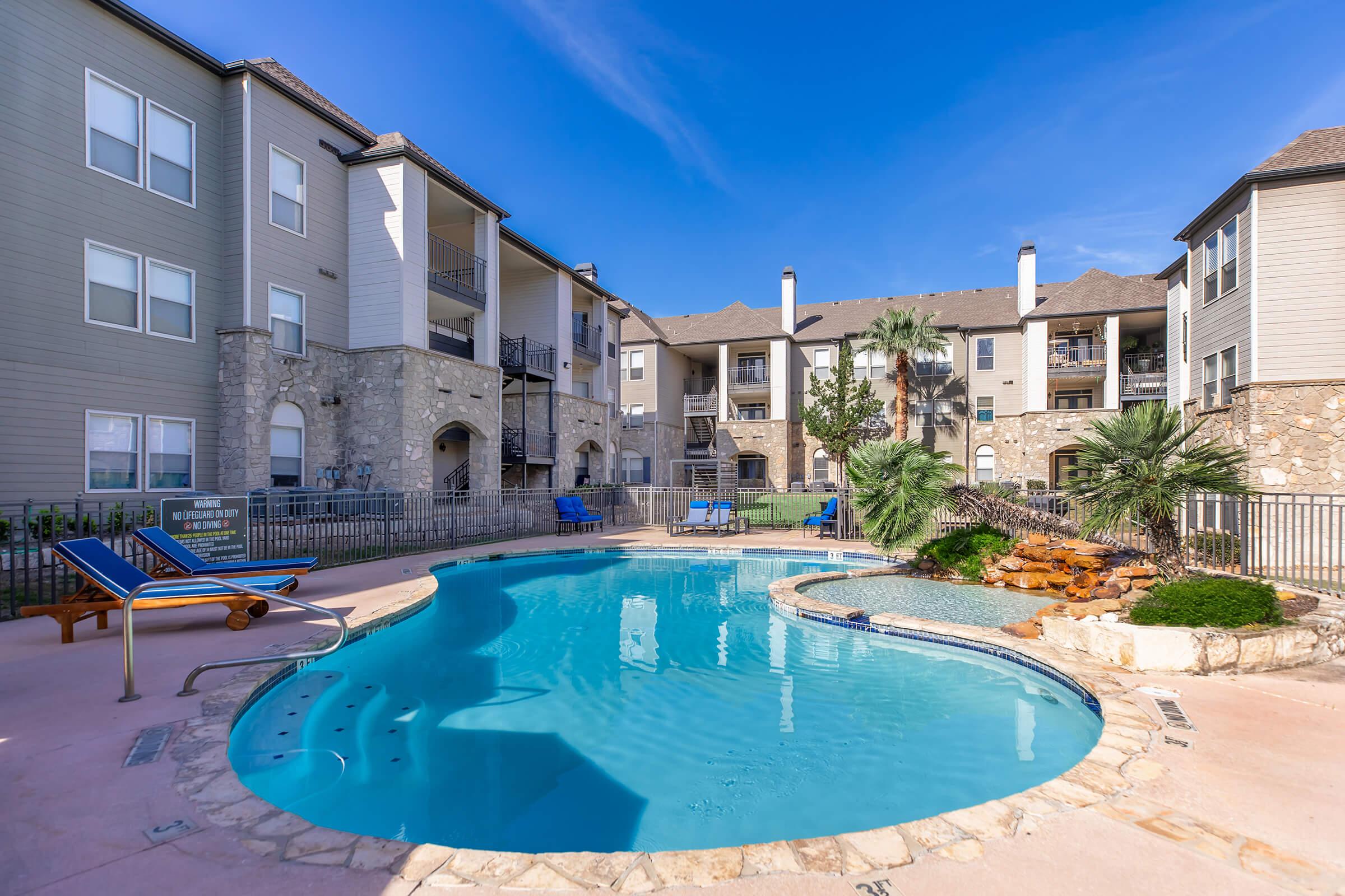 A spacious outdoor pool area surrounded by lounge chairs, palm trees, and landscaped rock features. The pool has a curved design with a waterfall feature and is located in the courtyard of a multi-story apartment complex with stone accents and balconies. Clear blue skies overhead.