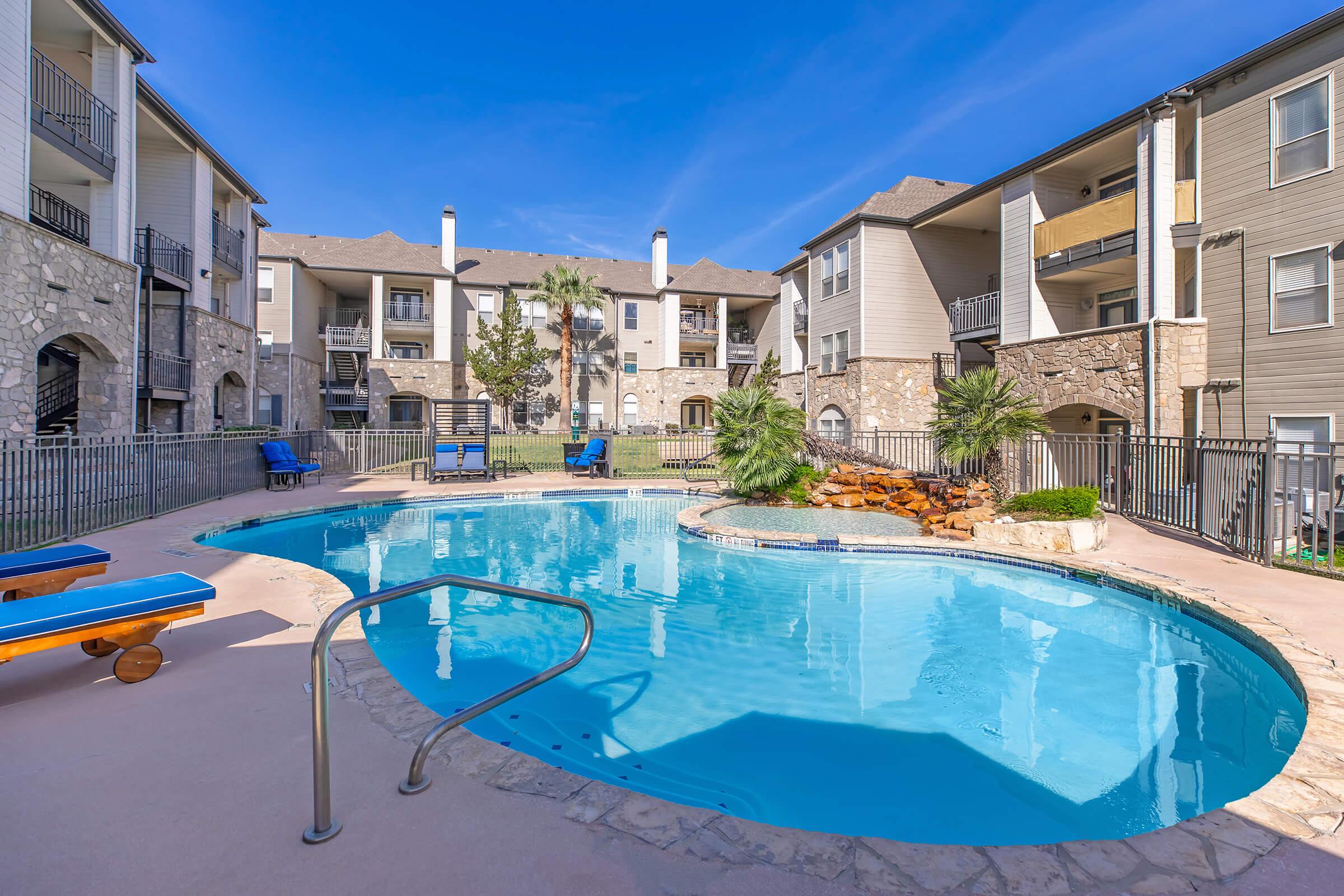 A resort-style swimming pool with a curved design, surrounded by lounge chairs and palm trees. The pool is located in a well-maintained apartment complex, featuring stone and wood architecture. Clear blue skies and bright sunlight enhance the inviting atmosphere.