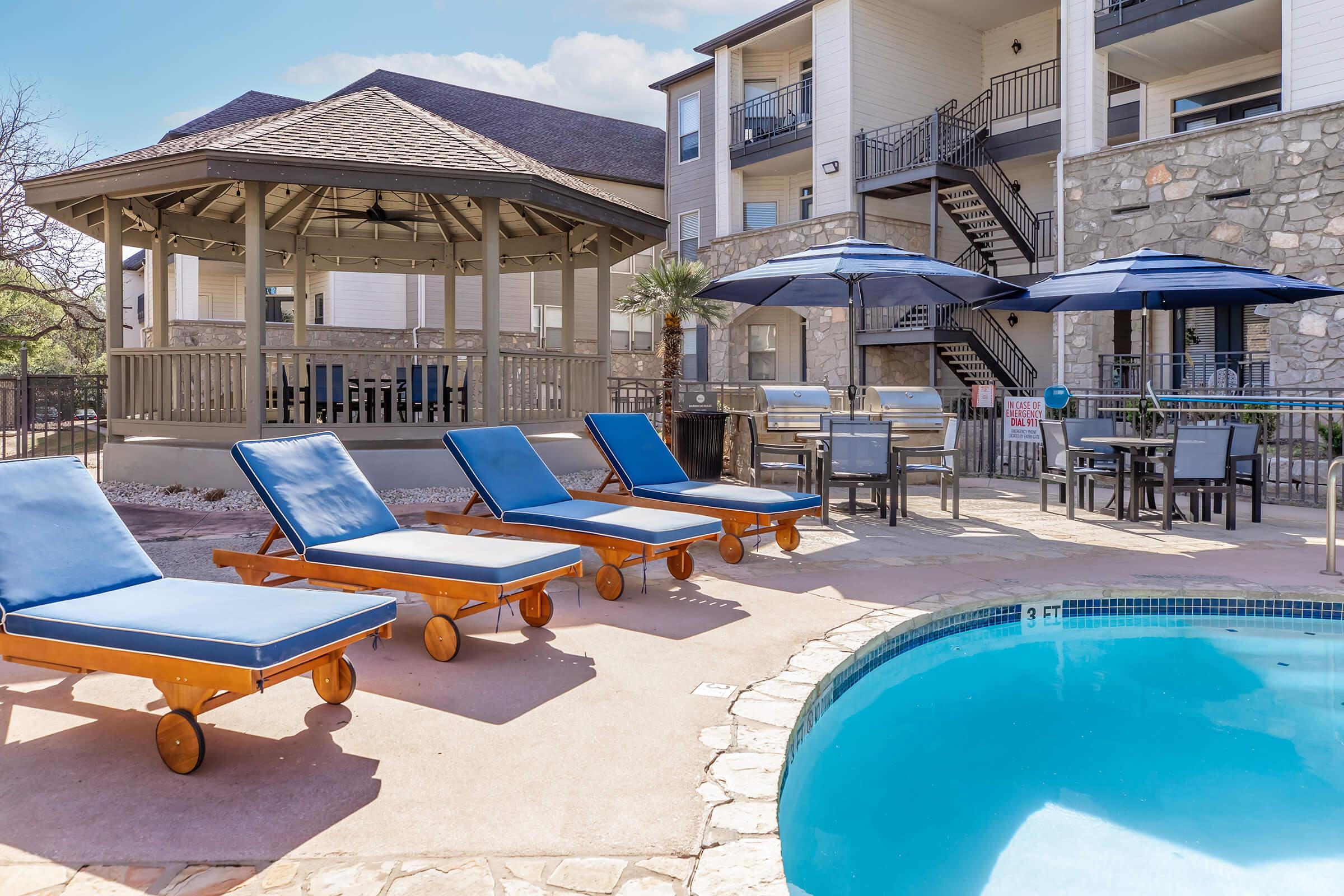 A sunny outdoor pool area featuring blue lounge chairs on a patio, a gazebo with seating, and umbrellas. The background includes a multi-story residential building with stairs and balconies. The scene is inviting and designed for relaxation.