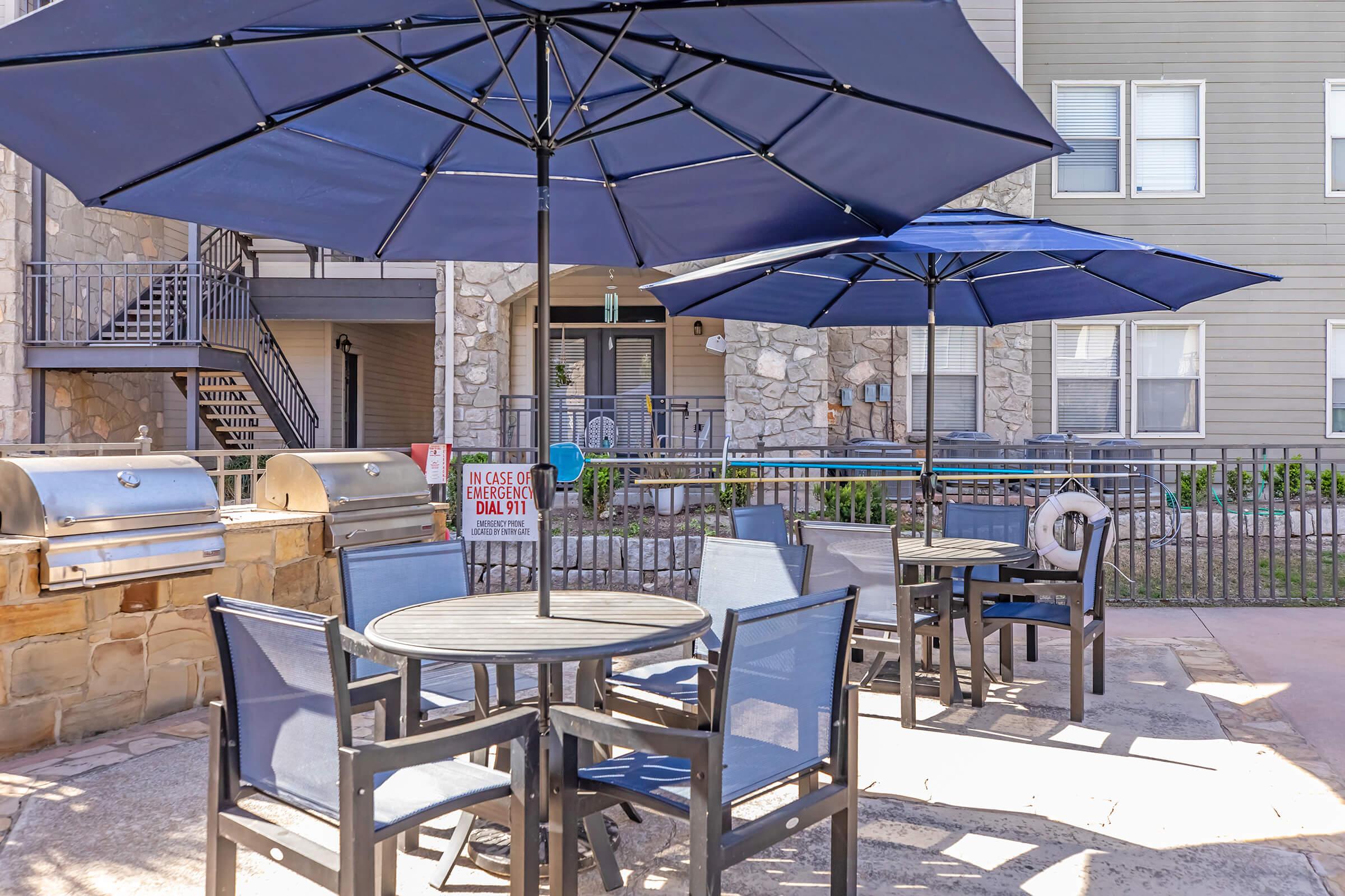 A patio area featuring several tables and chairs under large blue umbrellas. There is a grill nearby, and the background shows a multi-story apartment building with stone and siding exterior. A sign indicating emergency instructions is visible in the area.