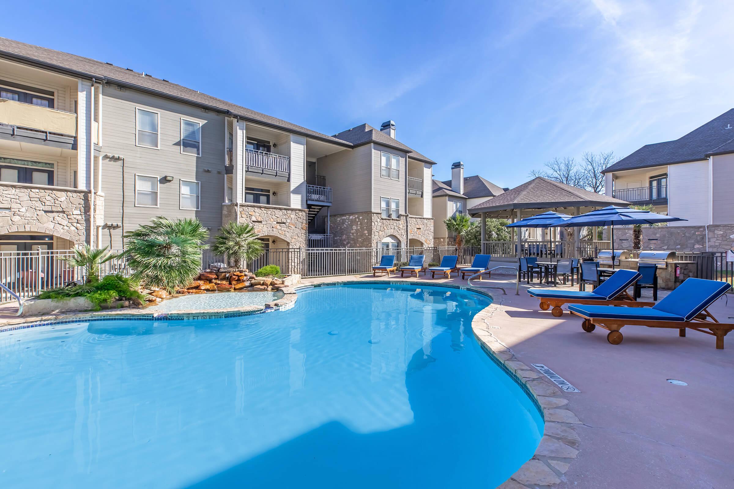 A sunny outdoor pool area featuring a clear blue swimming pool surrounded by lounge chairs and shaded seating. Apartment buildings in the background display a modern design with balconies. Lush landscaping adds a tropical feel to the serene atmosphere.