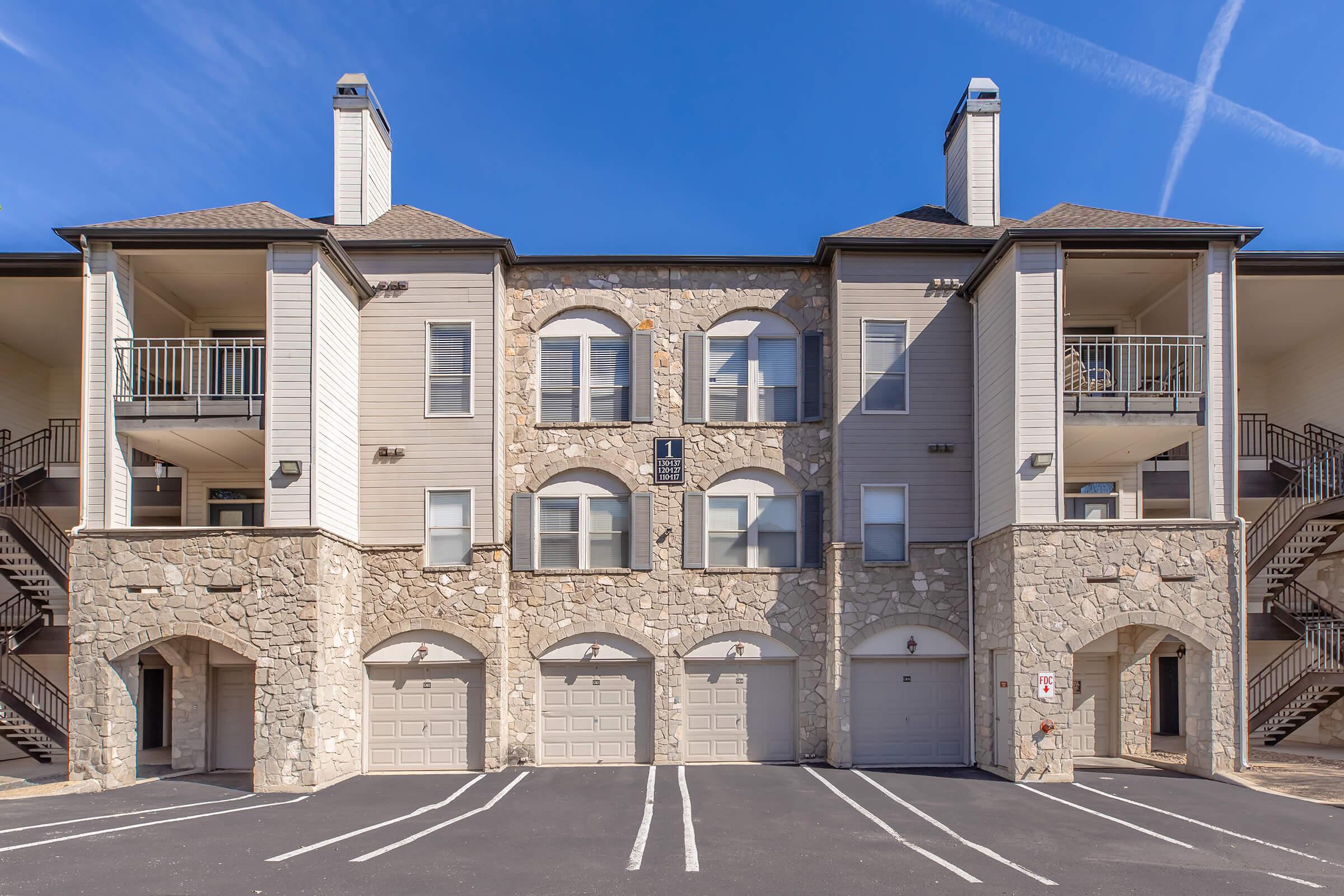 A two-story apartment building with a stone facade and beige siding. It features garages on the ground floor, balconies on the second floor, and a clear blue sky above. White parking lines indicate designated parking spaces in front of the building.
