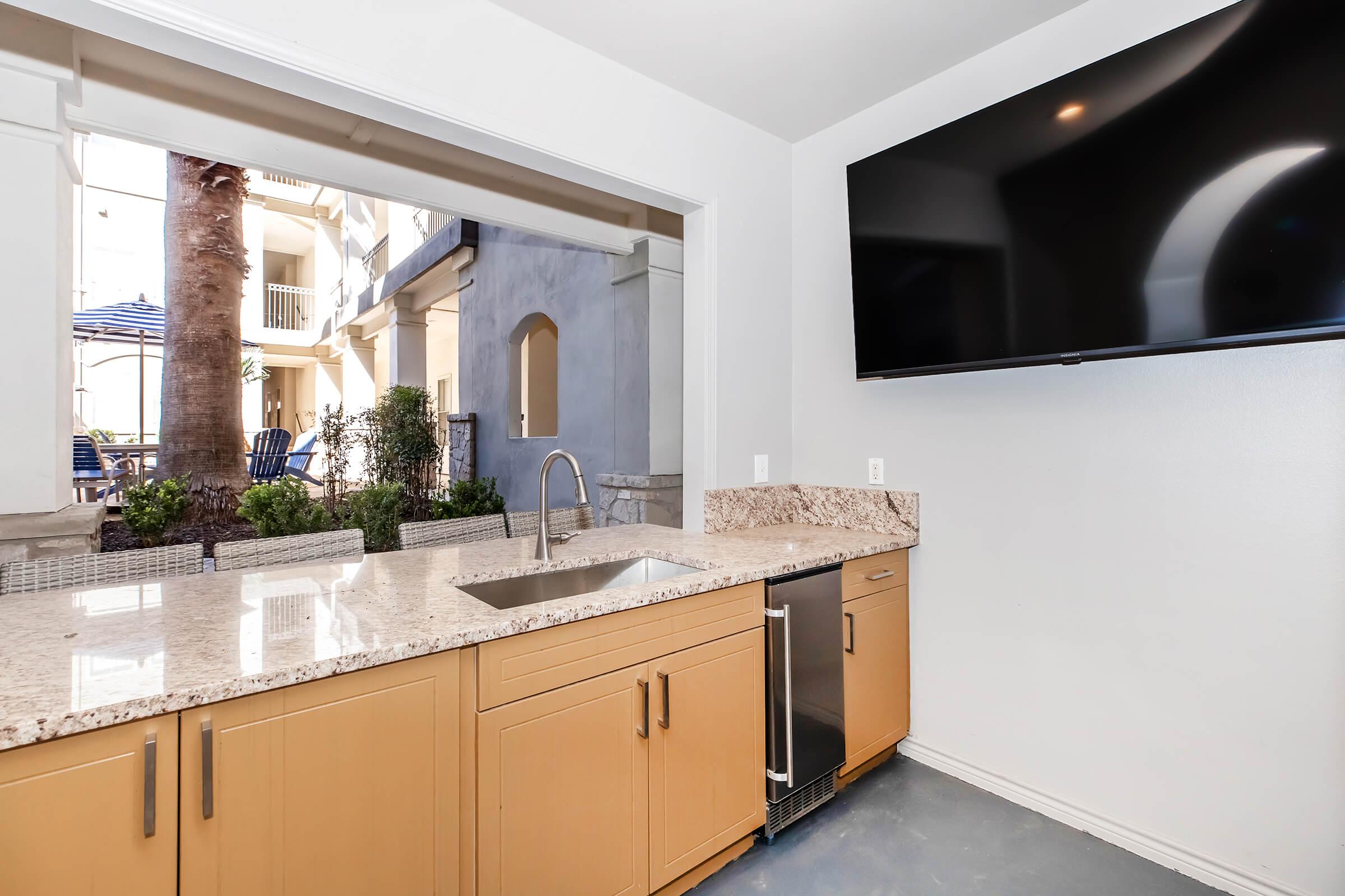 Modern kitchen space with sleek granite countertops, a stainless steel sink, and a refrigerator. A large wall-mounted flat-screen TV is visible. The area opens up to a courtyard featuring palm trees and outdoor seating, creating a bright and airy atmosphere.