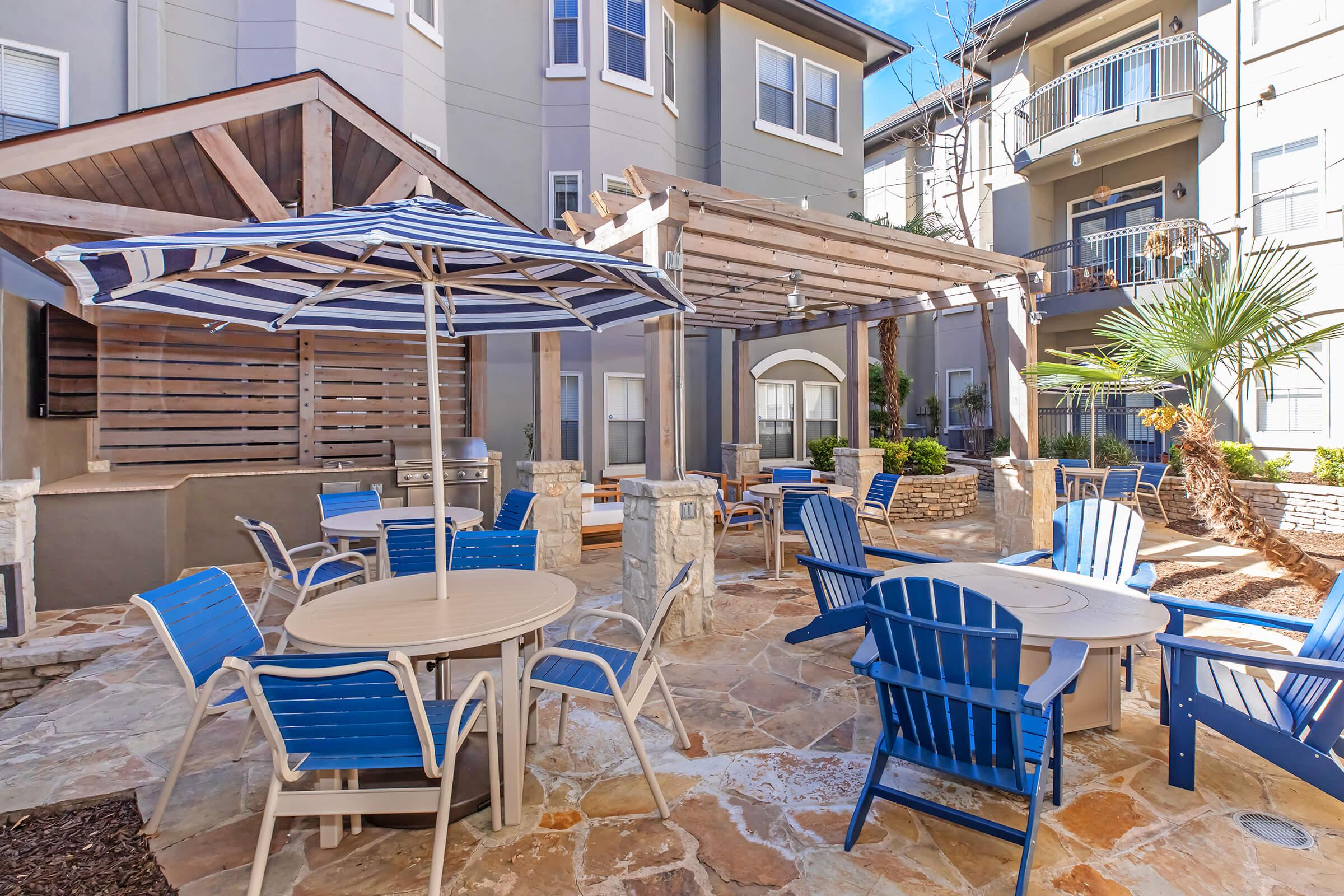 Outdoor patio area featuring stone flooring, several blue chairs, and tables. A large striped umbrella provides shade, and there are pergolas and palm trees in the background. The setting is well-lit with clear skies, creating a welcoming atmosphere for relaxation or social gatherings.