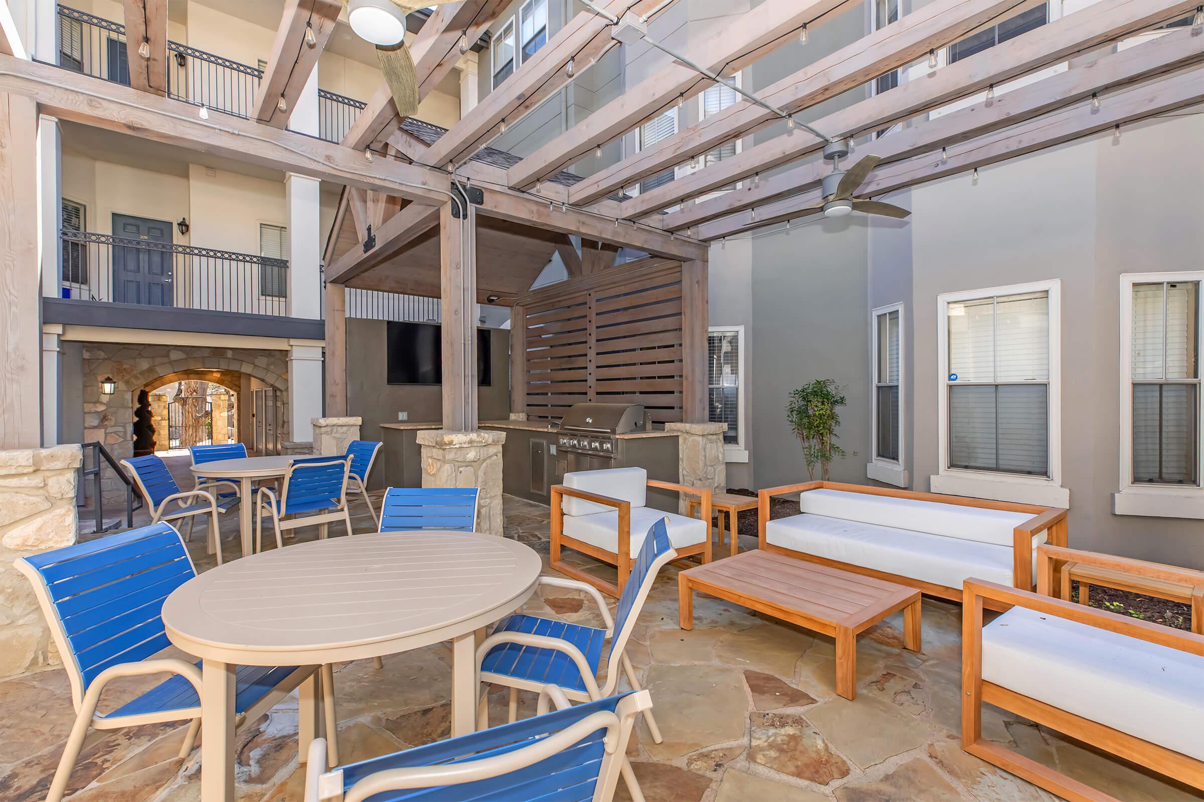 A cozy outdoor seating area featuring a round table with chairs, wooden benches, and a grill, all under a wooden pergola. The surrounding walls display a modern architectural style with large windows, creating a welcoming atmosphere for gatherings.