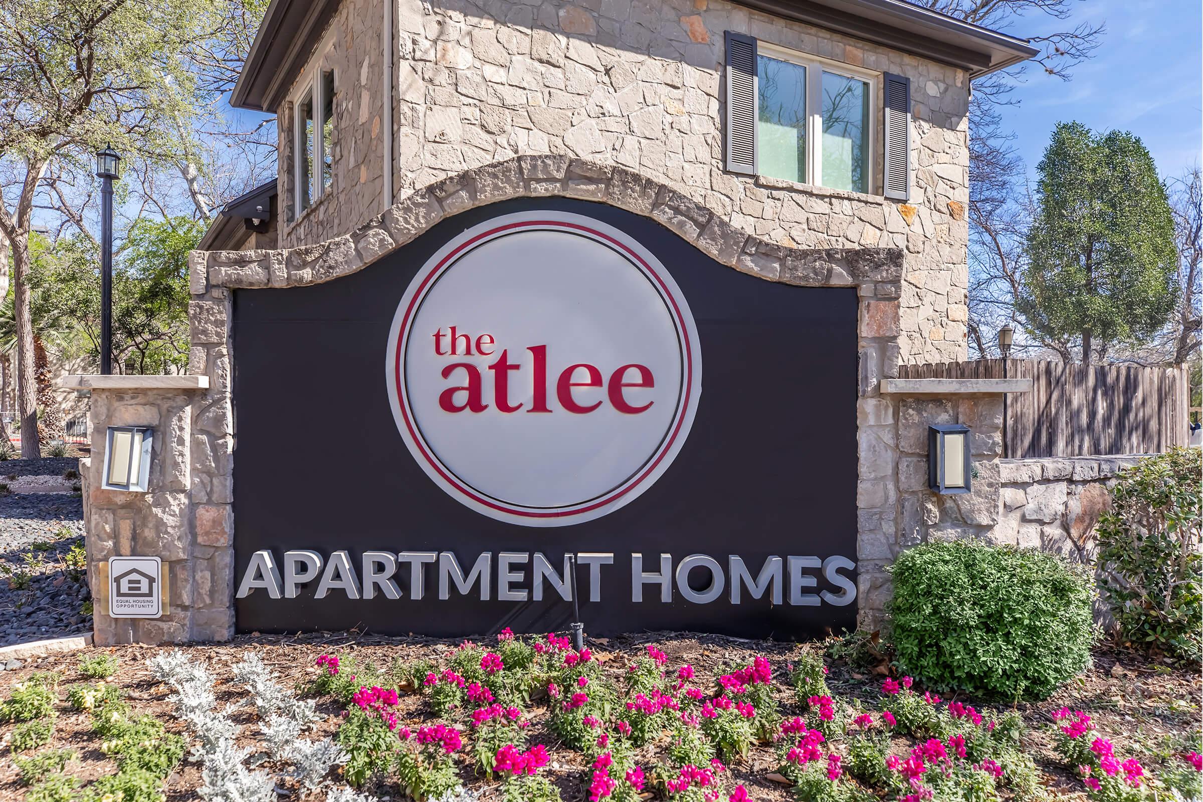 Sign for "The Atlee Apartment Homes" featuring a circular logo with the name prominently displayed. The sign is set against a stone wall with decorative landscaping, including pink flowers at the base.