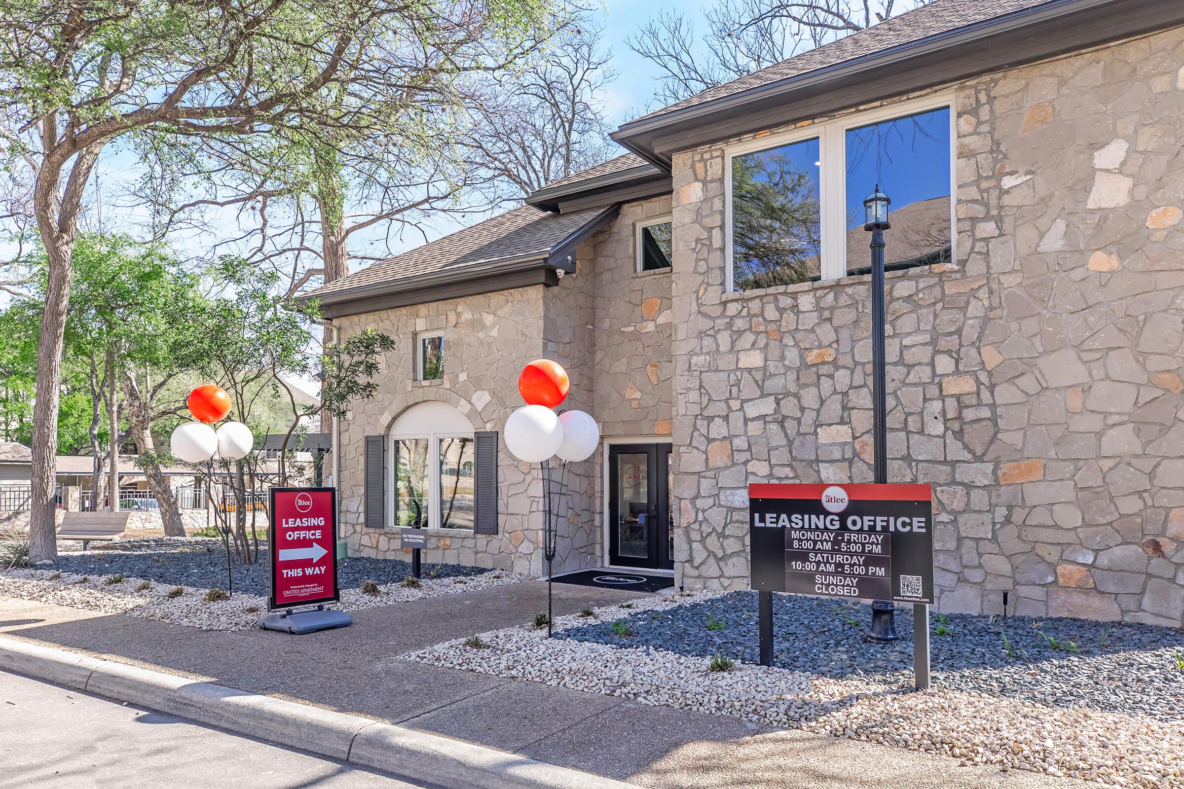 Leasing office building with a stone exterior and large windows. There are two red and white balloon decorations near the entrance, and signs indicating the office's hours. The surrounding area features trees and decorative gravel landscaping.
