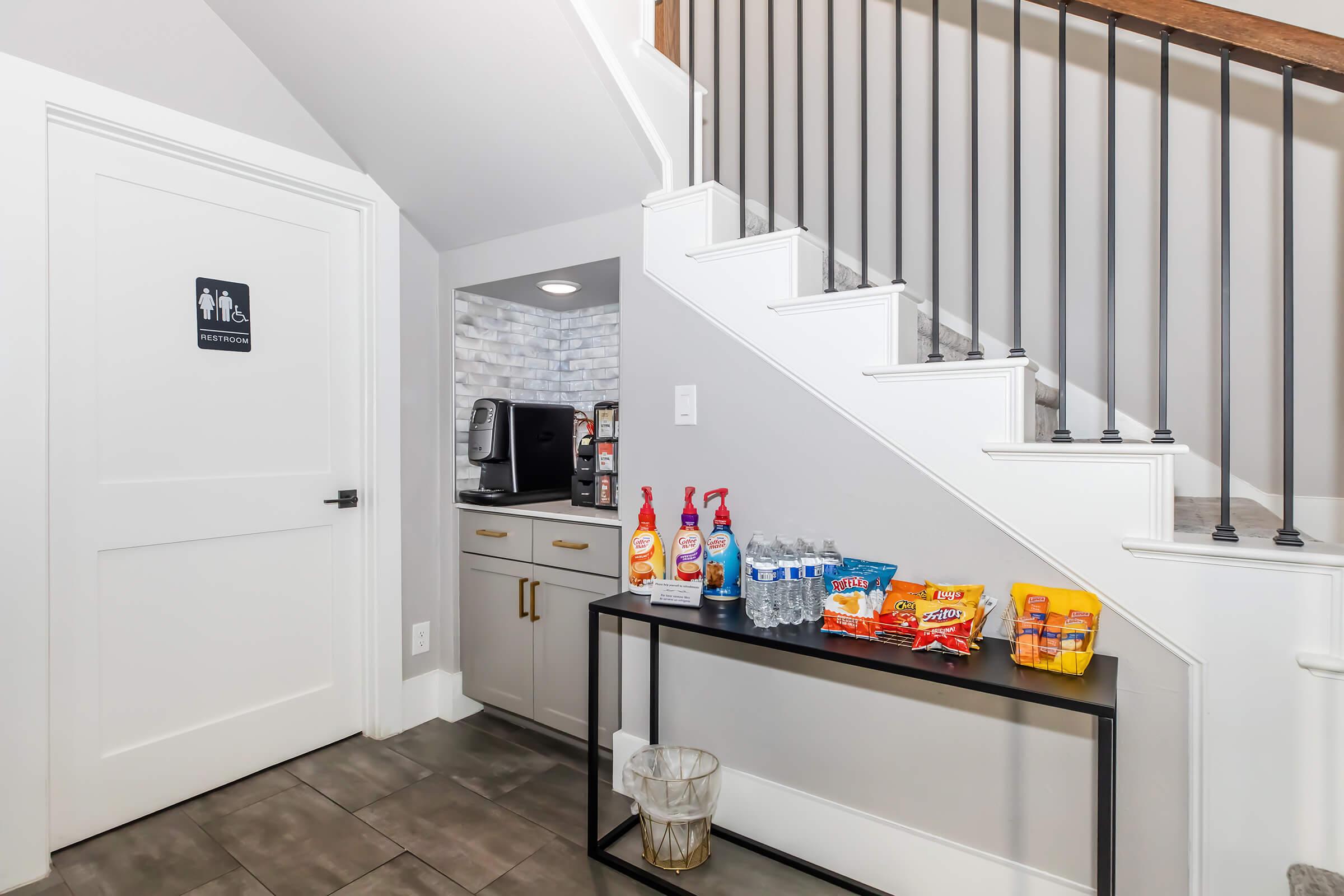 A well-lit interior space featuring a staircase, a door labeled "Restroom," and a small table. The table is stocked with various snacks, bottled water, and cleaning supplies. The wall has a modern design with gray tiles and a countertop area that includes a coffee maker.