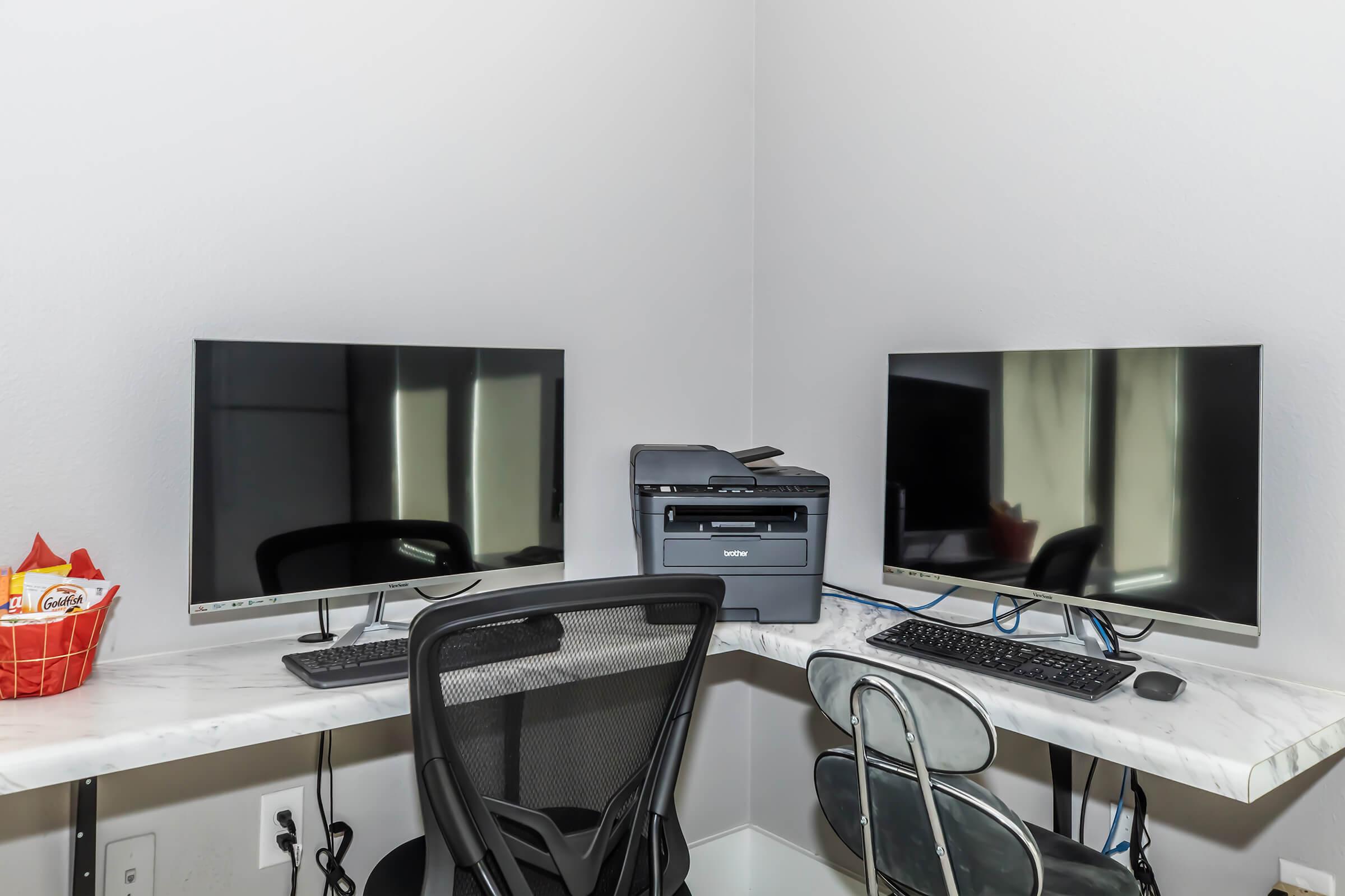 Two computer monitors with black screens sit on a marble-top desk, one accompanied by a printer in the middle. A stylish office chair is positioned at each monitor. A basket of snacks is visible in the corner. The room has soft lighting and neutral-colored walls.