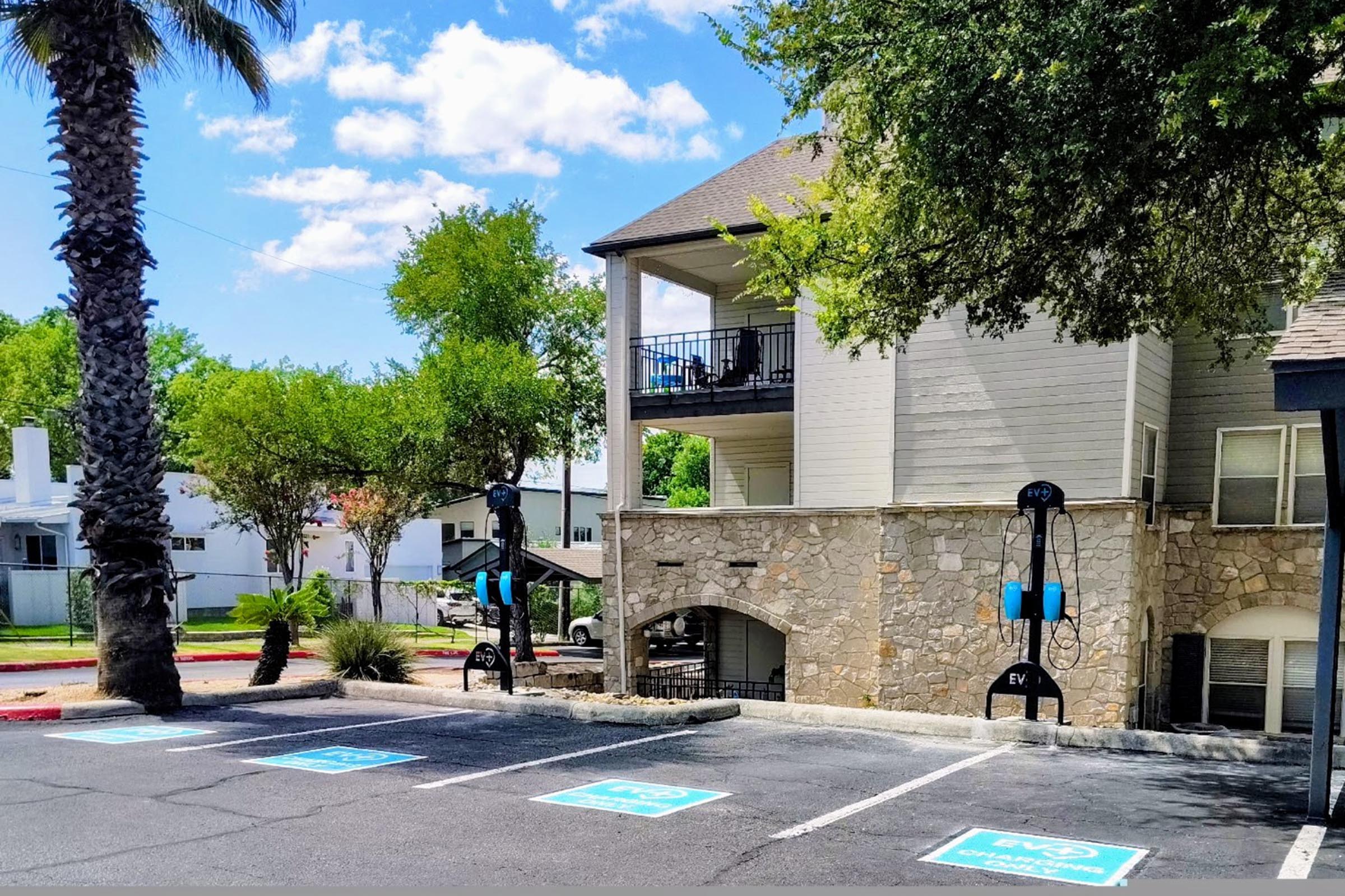 A residential building with a stone façade and balconies is seen under a clear blue sky with clouds. In the foreground, there are designated electric vehicle charging stations marked with blue signs in a parking area surrounded by greenery and palm trees.