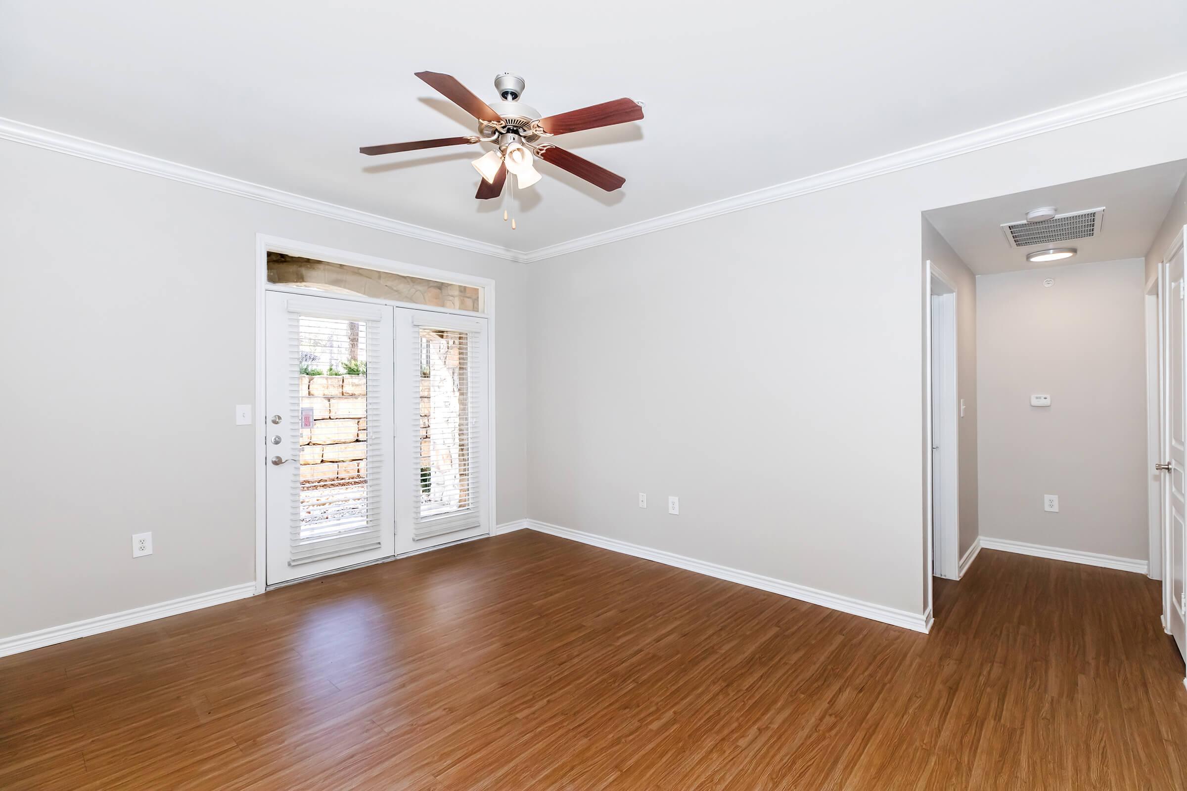 Interior view of a spacious room featuring hardwood-style flooring, light gray walls, and a ceiling fan with wooden blades. There are two sets of French doors leading outside, with blinds partially closed. A hallway is visible on the right side of the image, suggesting access to other areas.