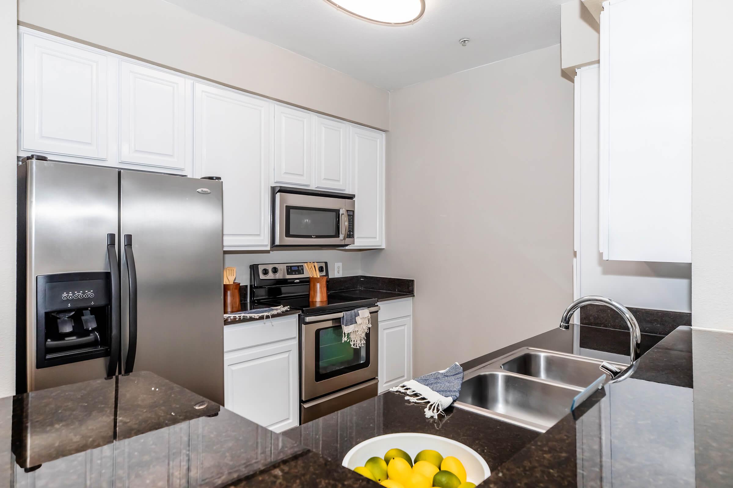 Modern kitchen featuring stainless steel appliances, including a refrigerator and microwave. The countertops are dark granite with a bowl of lemons and limes. White cabinetry provides a clean look, and the space is well-lit with a ceiling fixture.