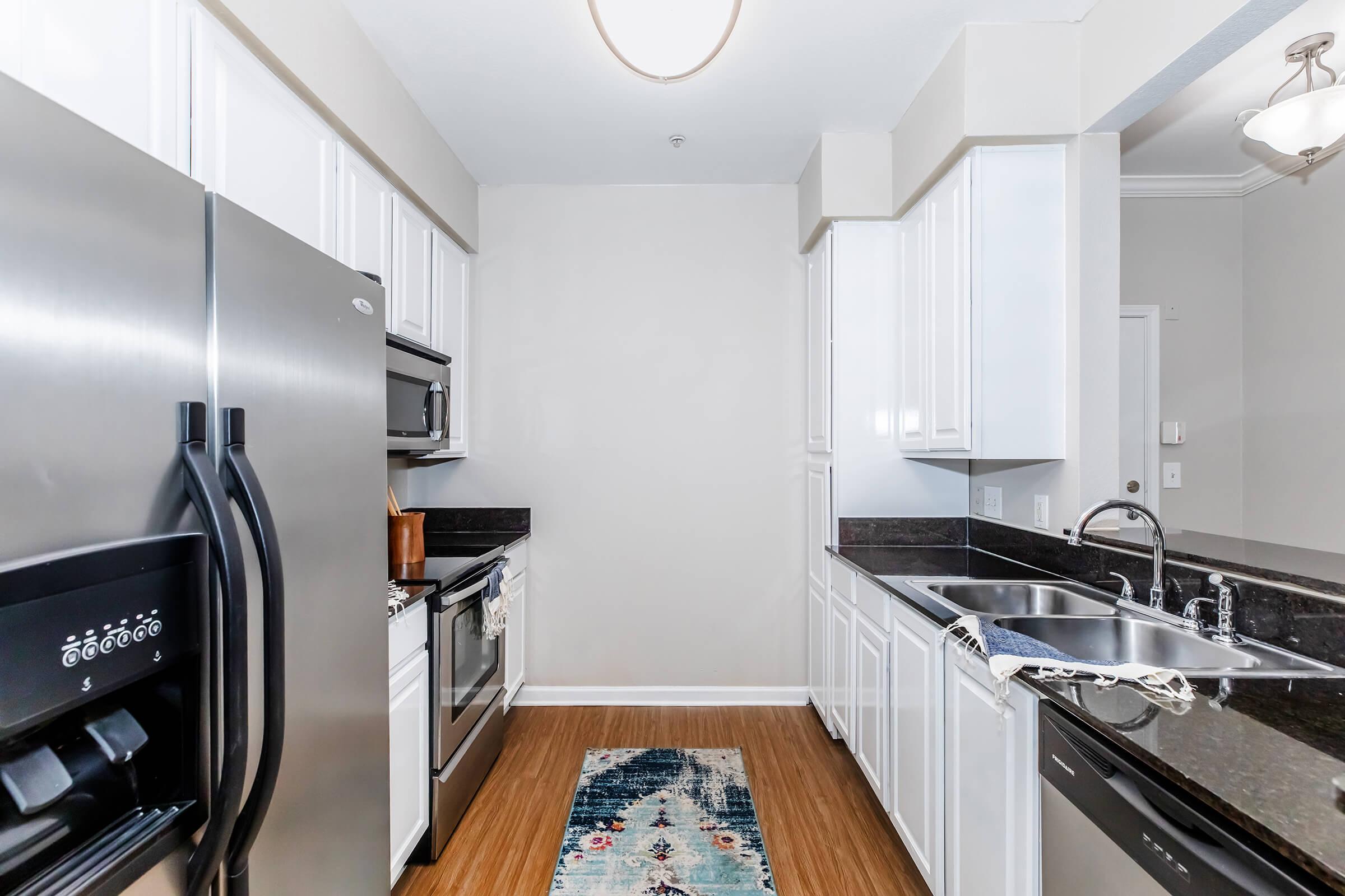 Modern kitchen with stainless steel appliances, including a refrigerator, oven, and microwave. Features white cabinetry, black countertops, and a hardwood floor. A blue patterned rug is placed in the center, adding a touch of color to the space. The kitchen is well-lit with a ceiling light fixture.
