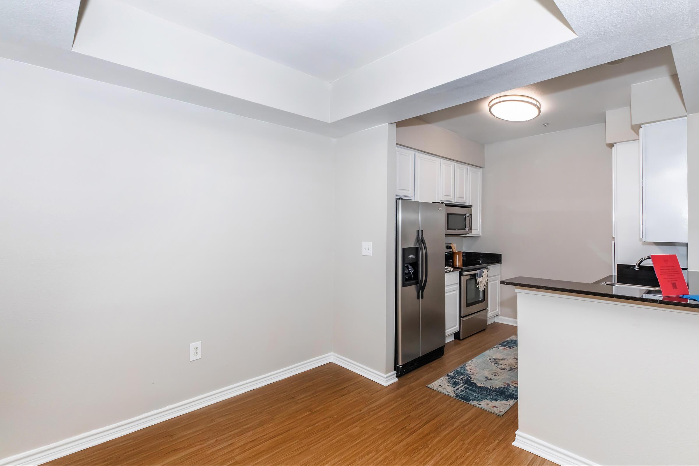 Modern kitchen area featuring stainless steel appliances, including a refrigerator and microwave, with dark countertops. The space is well-lit with overhead lighting and has a light-colored wall. A patterned rug is visible on the floor, adding a touch of decor to the area.