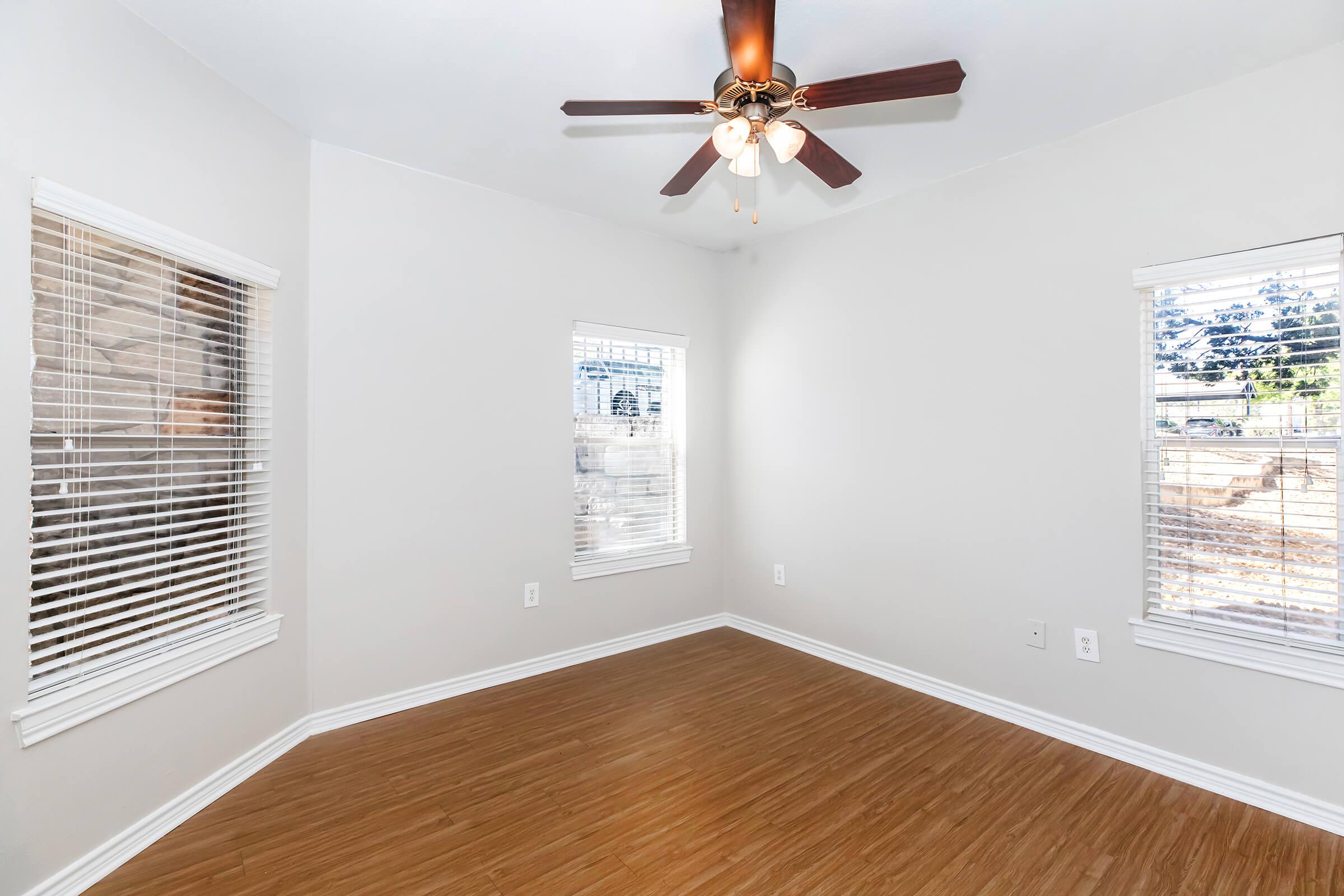 A well-lit empty room featuring light-colored walls and wooden flooring. It has two windows with blinds allowing natural light in, and a ceiling fan with light fixtures in the center. The overall atmosphere is clean and inviting, suitable for various uses such as a living area or office space.