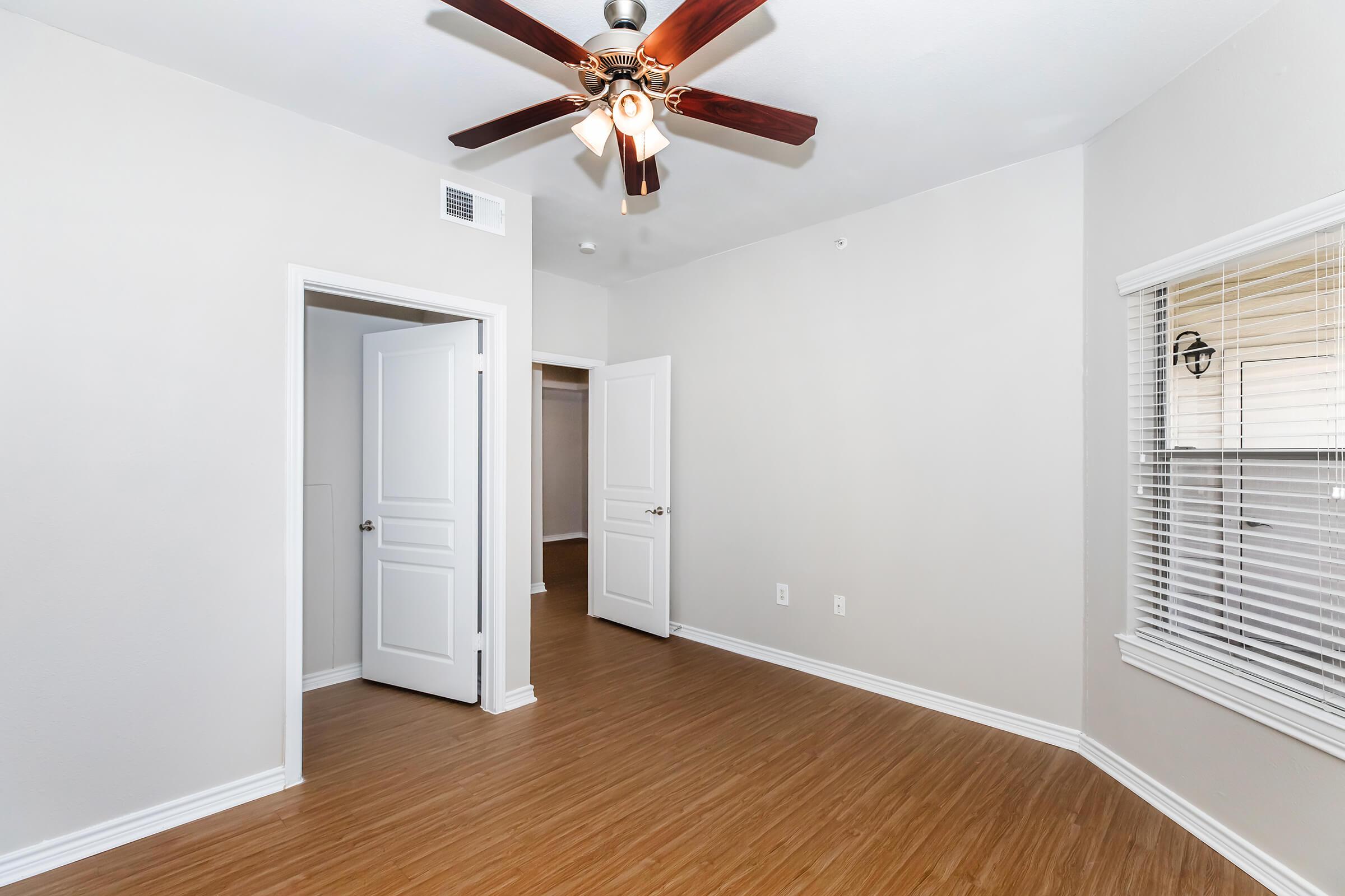 Interior of a room featuring light-colored walls and a ceiling fan with wooden blades. There are two white doors, one leading to a closet and another to the hallway. The flooring is bamboo-style, and there is a window with blinds providing natural light. The overall space appears bright and inviting.