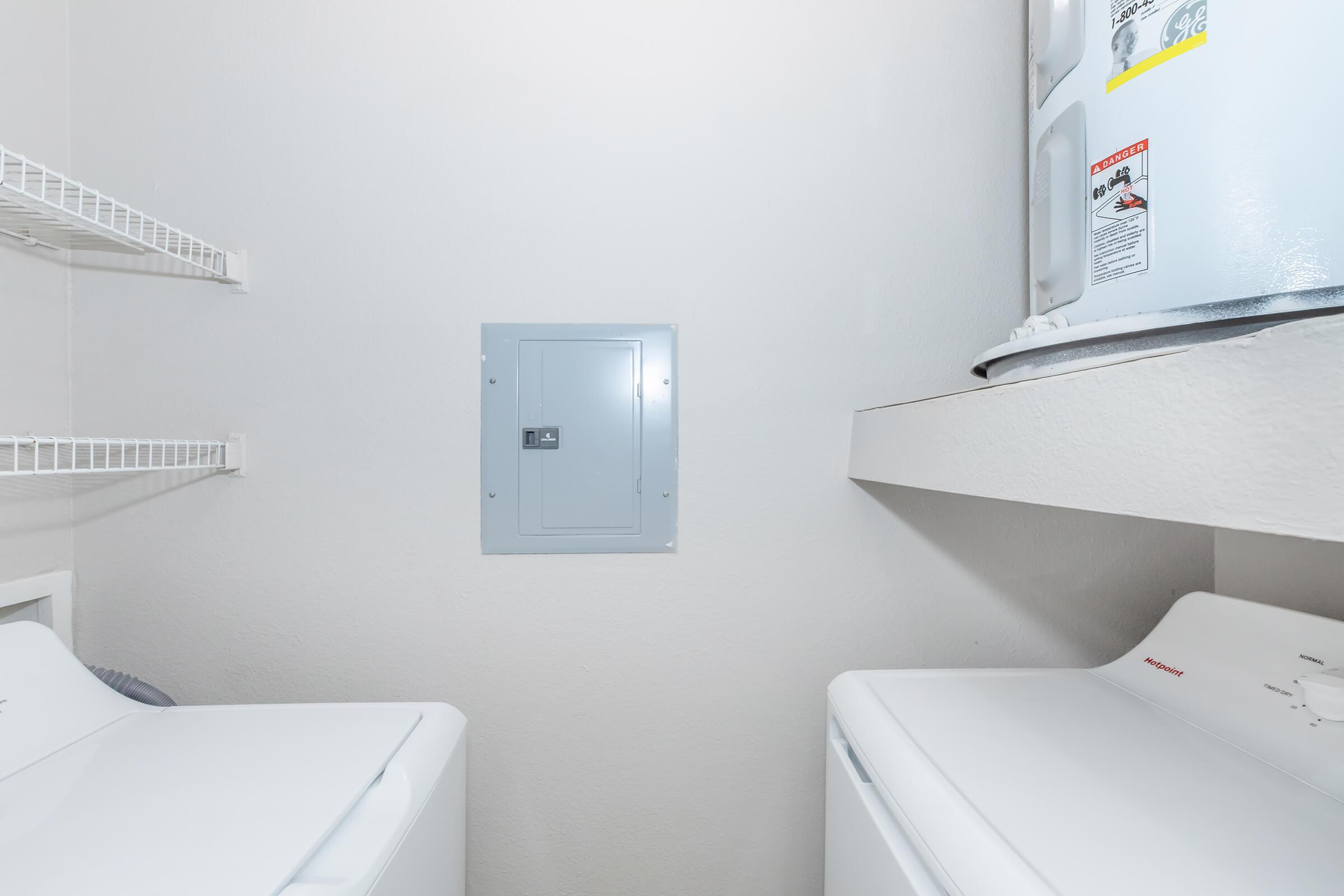 A laundry room with a washing machine on the left and a dryer on the right. There are white shelves on the wall for storage, and a gray electrical panel mounted on the wall. A water heater is visible in the corner, providing utility space in a bright, clean environment.