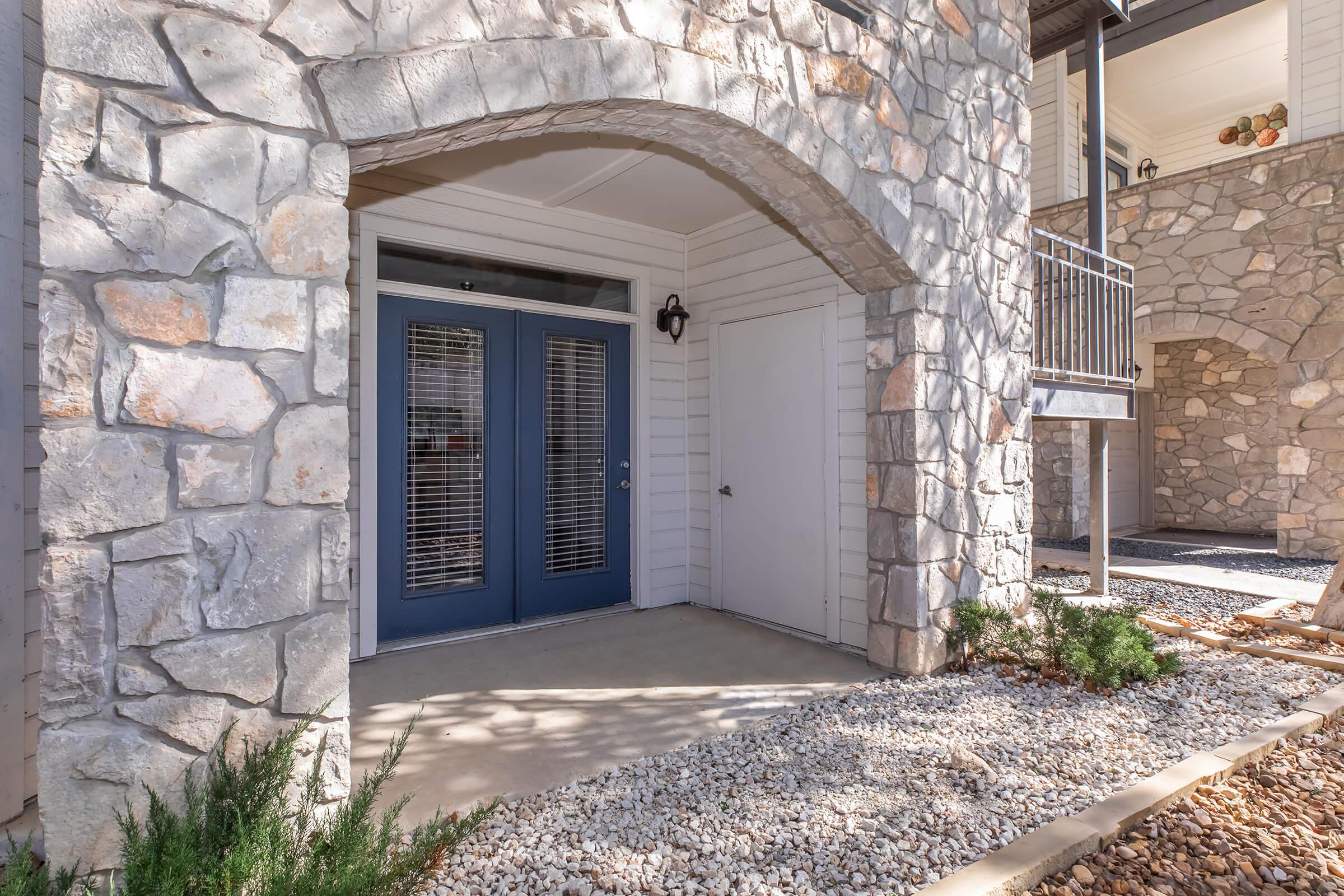 A stone-textured exterior reveals a welcoming entrance with two blue double doors and decorative shutters. The floor is concrete with a small garden area featuring rocks and low shrubs. A balcony is seen in the background, adding to the architectural charm.