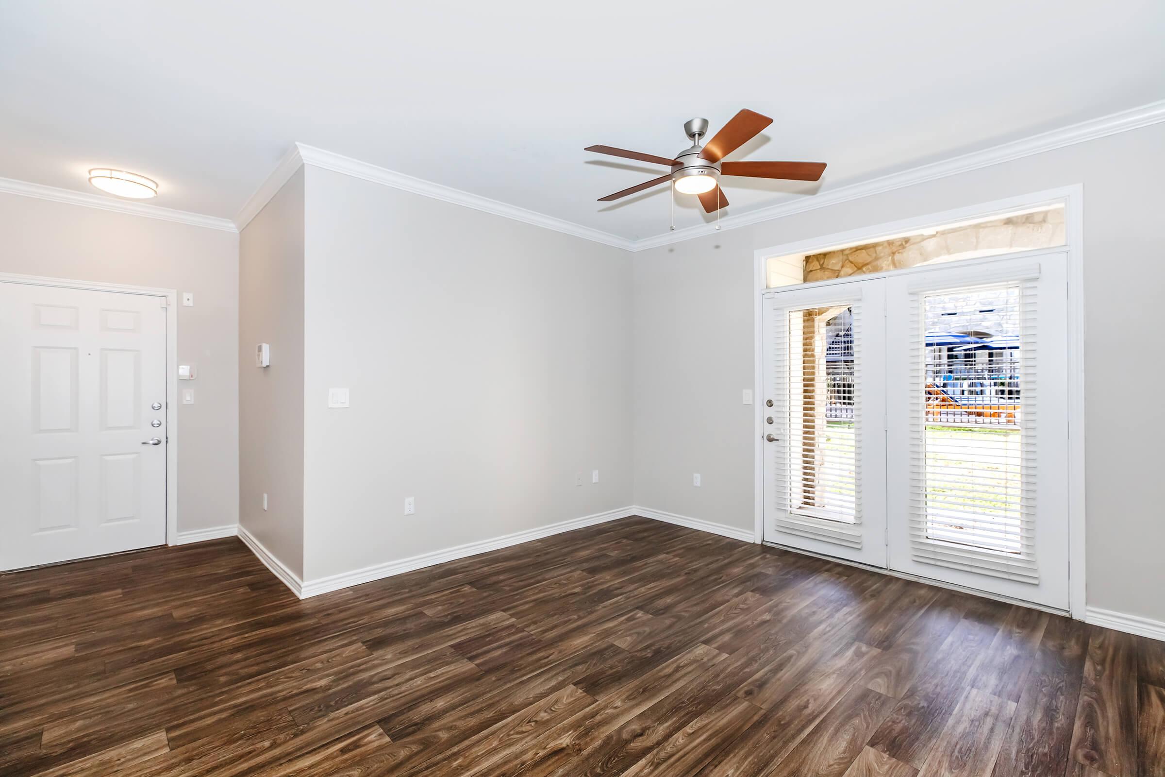 Bright and spacious living room with wood-style flooring, ceiling fan, and large windows. Features an entrance door on the left and double French doors leading outside to a patio or balcony. Walls are painted in a light neutral color, enhancing the open feel of the space.