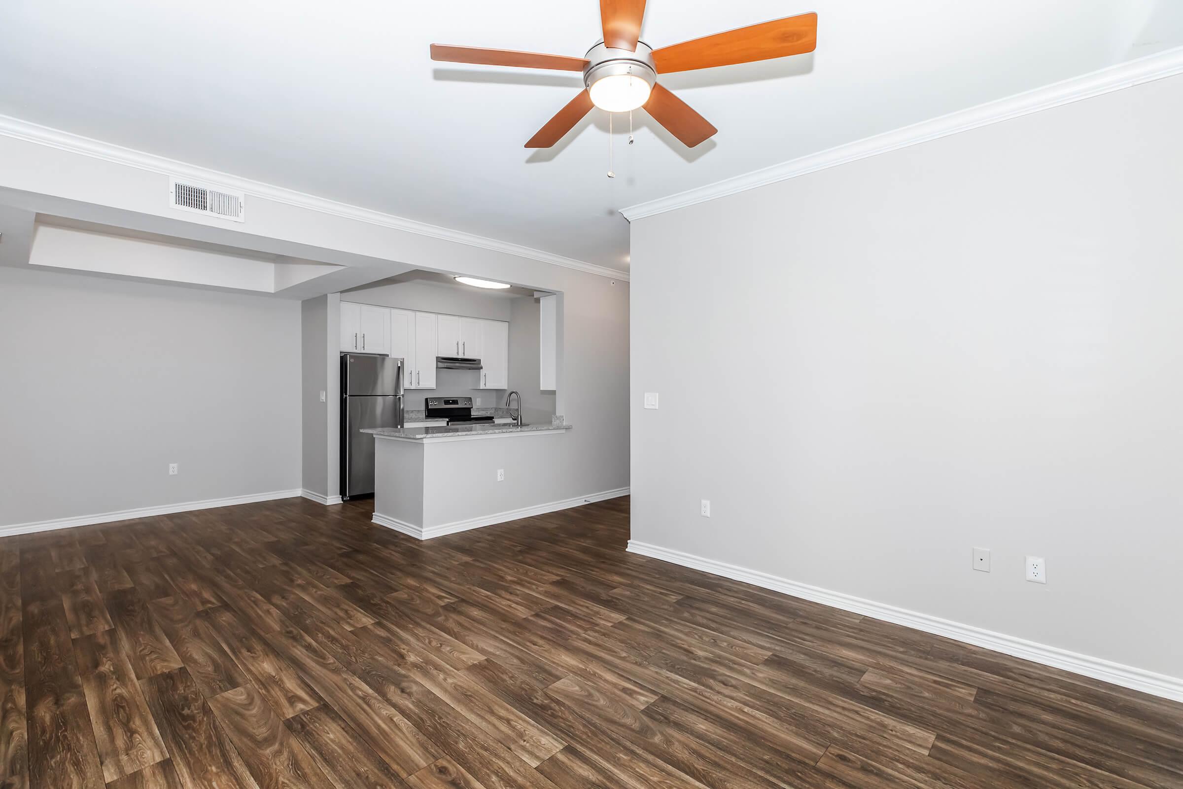 A modern, spacious living area featuring hardwood flooring, a ceiling fan, and neutral-colored walls. In the background, a kitchen with stainless steel appliances is visible. The layout is open and well-lit, creating a welcoming atmosphere.