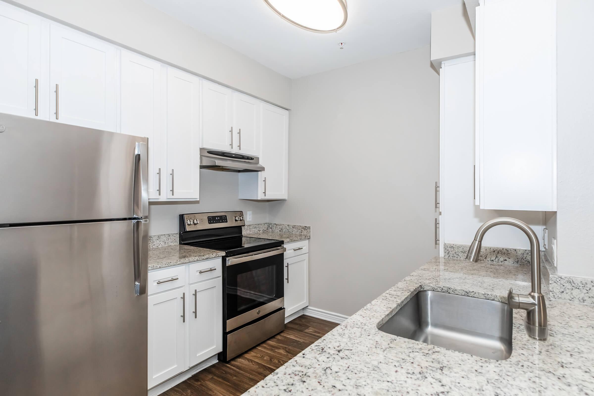 Modern kitchen featuring stainless steel appliances, including a refrigerator and an oven, granite countertops, and white cabinetry. The space has a sleek design with a light color palette and a single-basin sink, creating a clean and contemporary look.