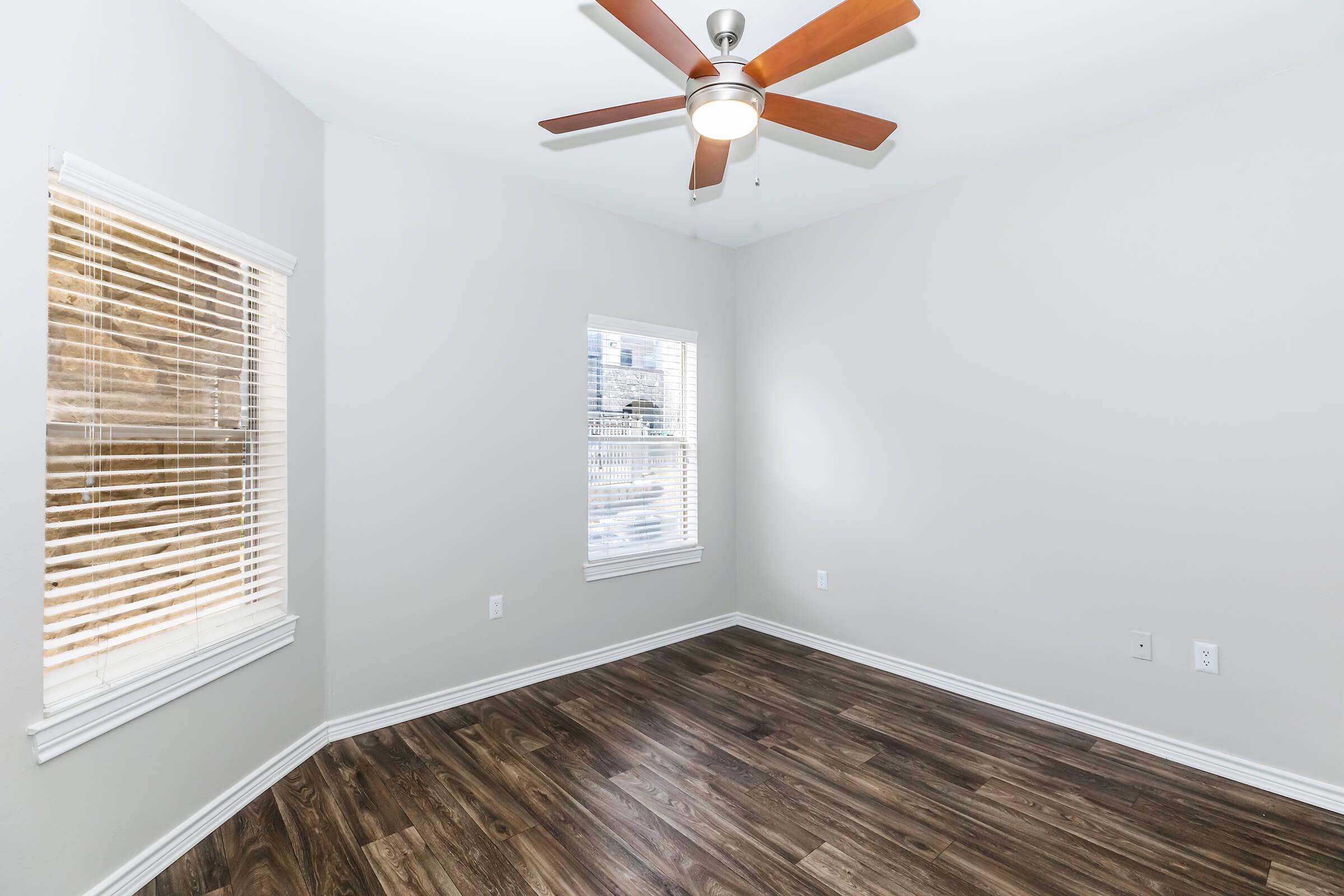 A bright, empty room with light gray walls and wooden floors. Two windows with blinds allow natural light to enter, and a ceiling fan with wooden blades is mounted in the center. The room appears spacious and ready for furnishing.