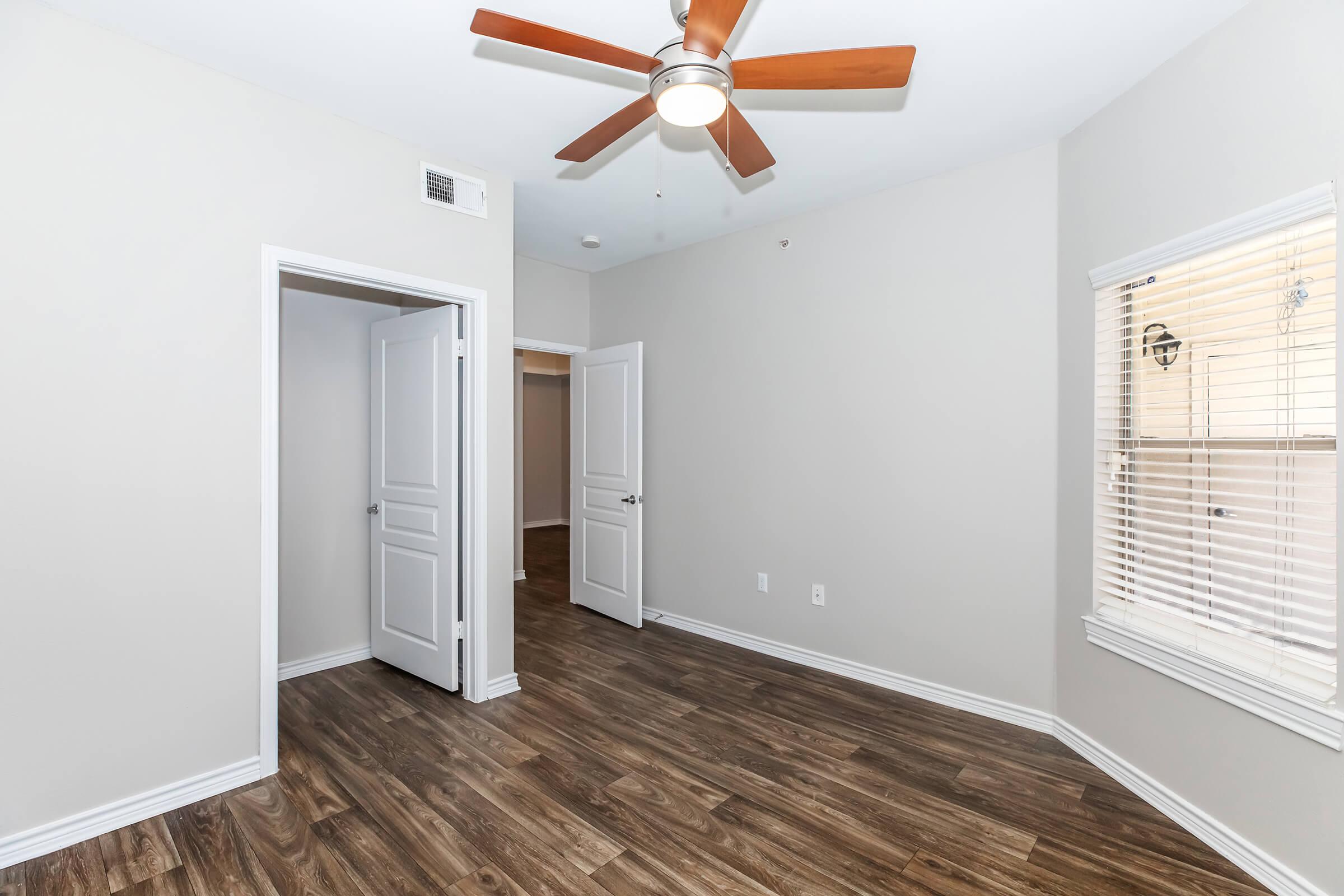 Empty room featuring light-colored walls, a ceiling fan, and wood-like flooring. Two white doors are visible, one leading to a closet. A window with blinds allows natural light to enter, illuminating the space.