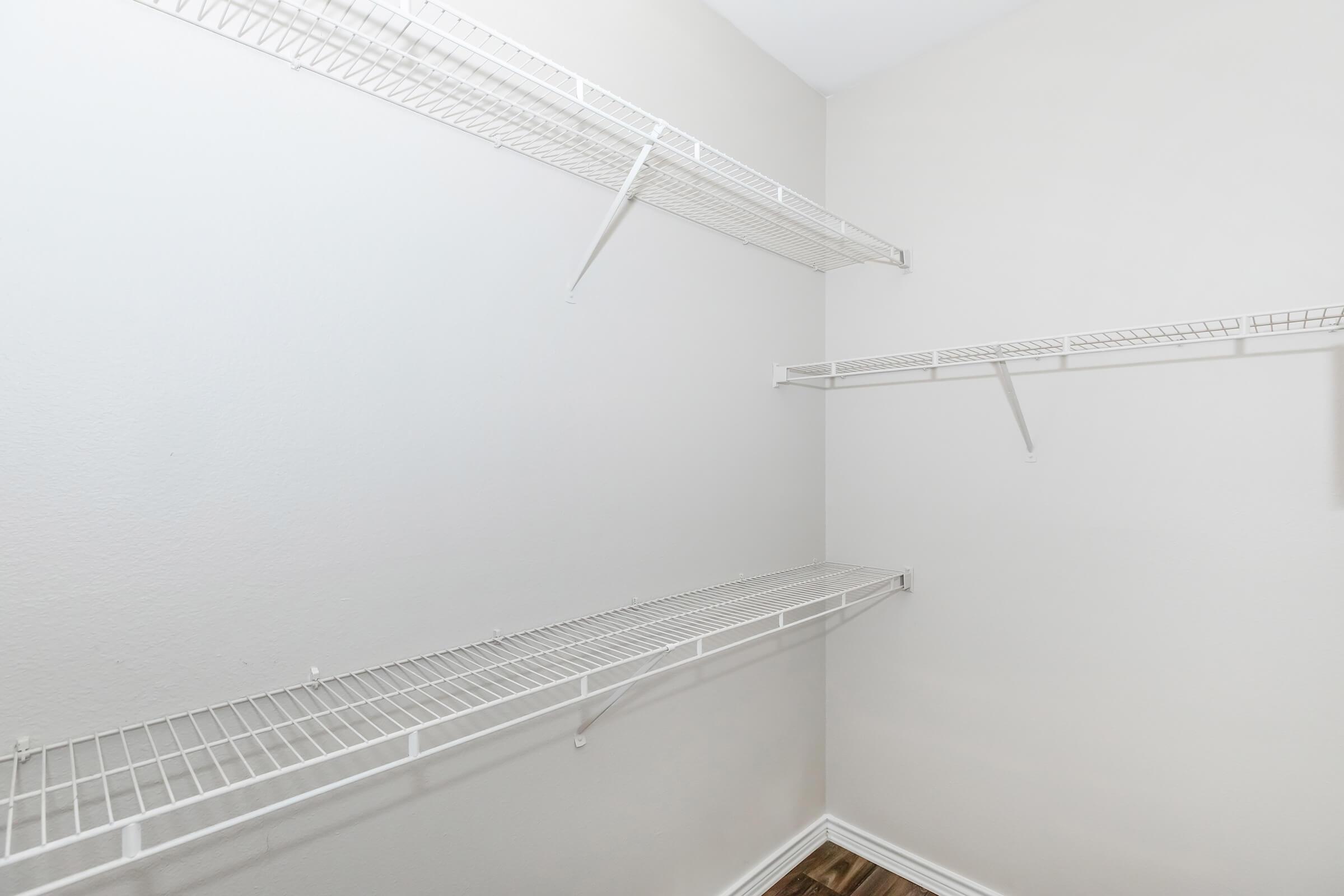 Empty closet space with white wire shelves attached to light-colored walls, featuring a wooden floor. The shelves are arranged at different heights, providing storage options for various items.
