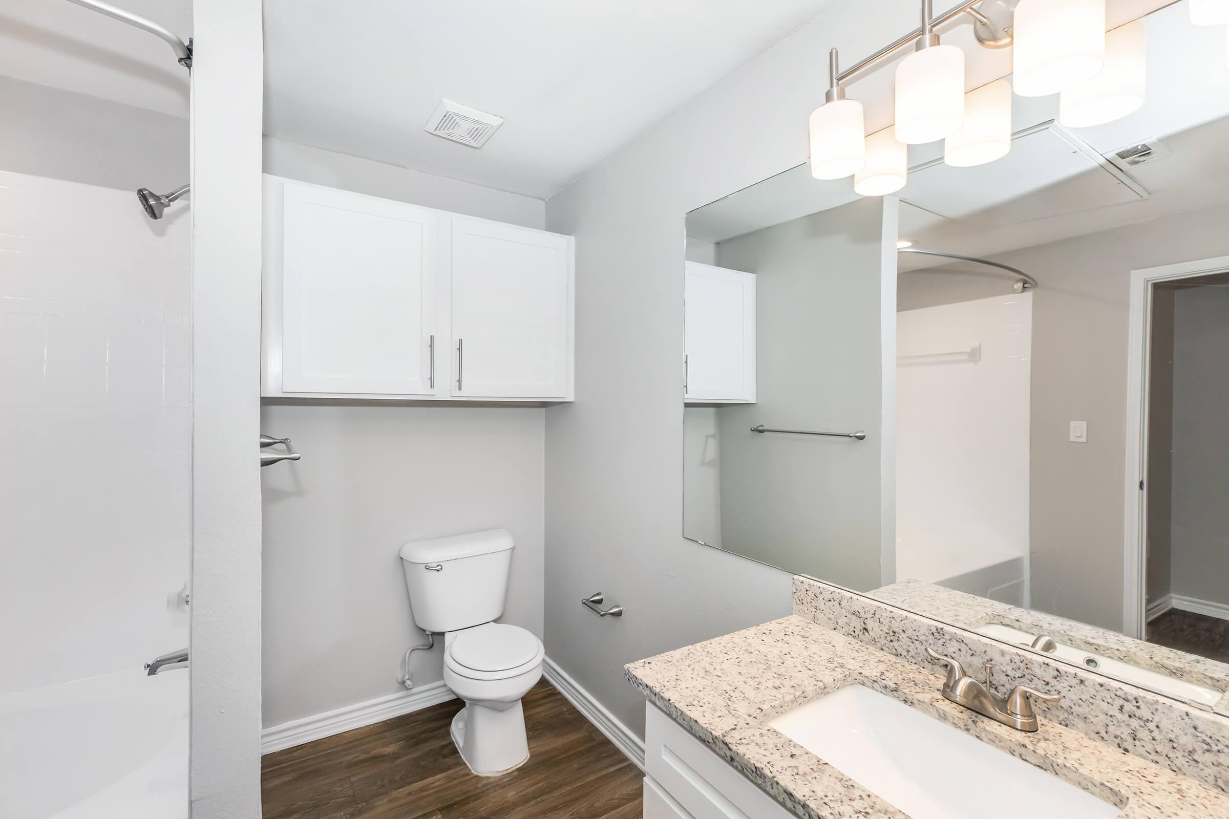 A modern bathroom featuring a white toilet, a granite countertop with a sink, a large mirror, and overhead lighting. The walls are painted in a light gray color, and there is a shower area with a white curtain. Cabinets provide storage space, and the floor is made of wood laminate.