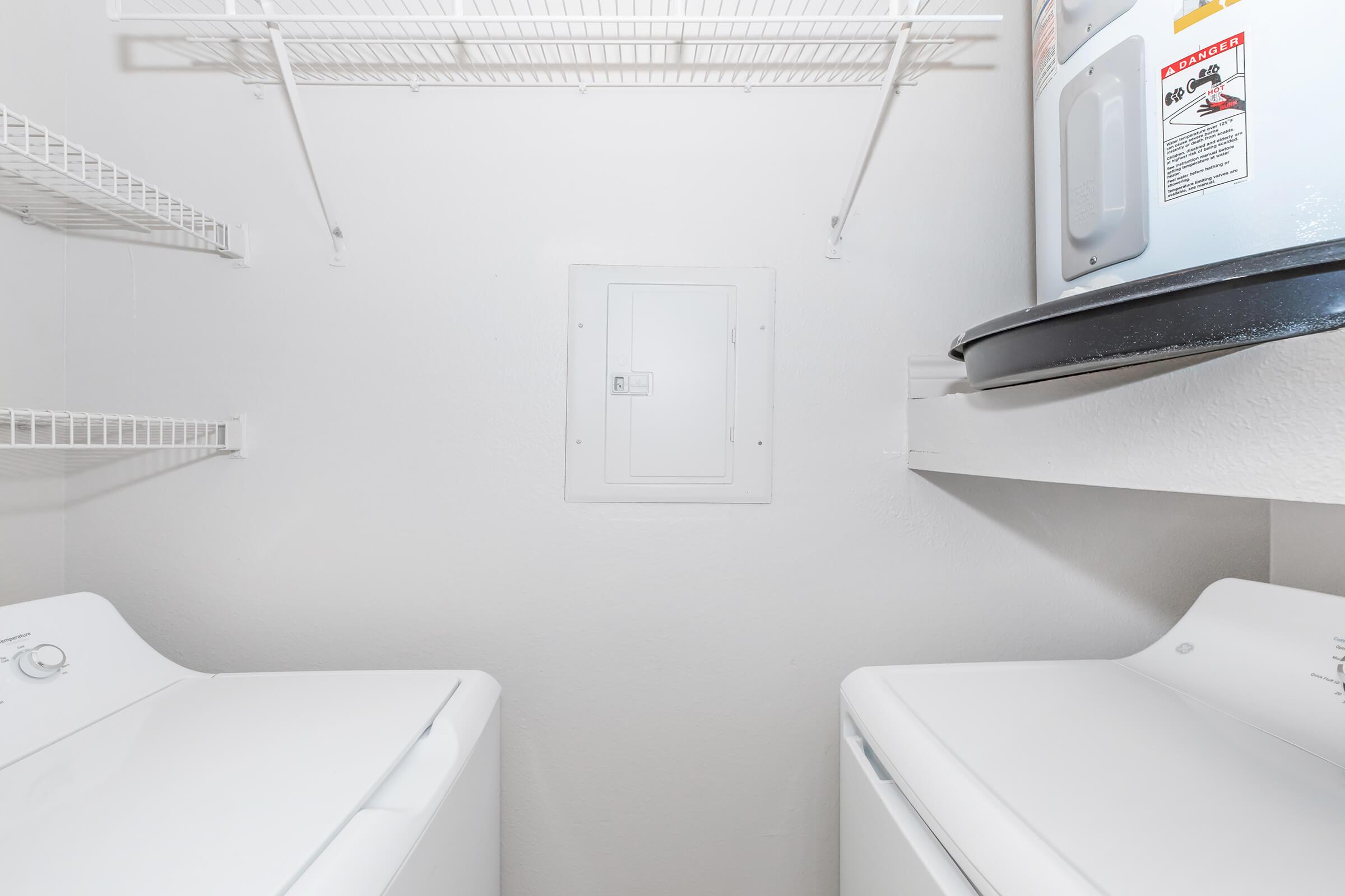 A clean, small laundry room featuring two white washing machines, white wire shelving on the walls, and a water heater in the corner. The walls are painted light colors, creating a bright and spacious appearance.