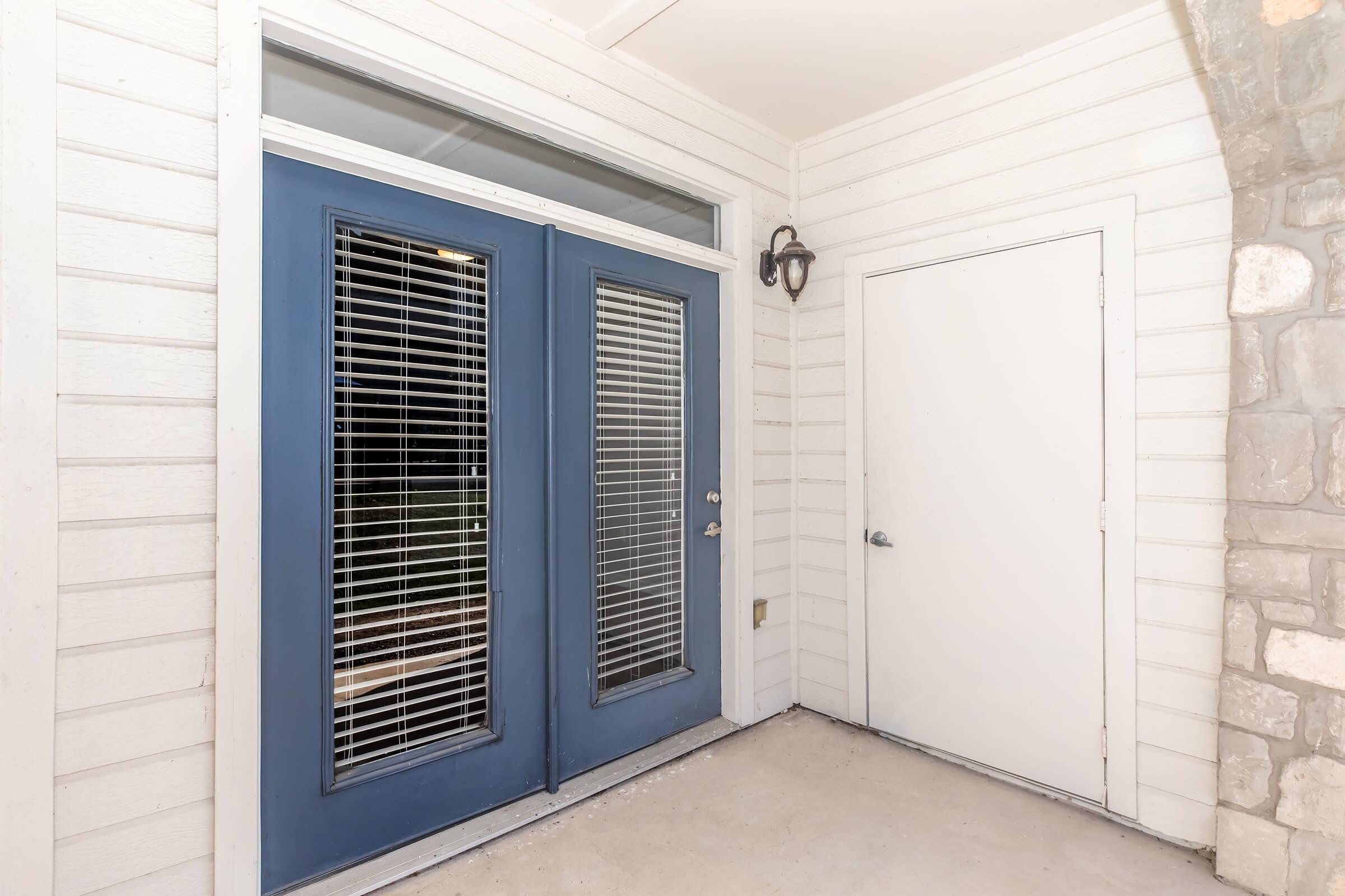 A well-lit entryway featuring double blue doors with vertical slat windows, flanked by a single white door. The surrounding wall is made of white wooden panels and stone, providing a clean and modern look. A wall-mounted light fixture is positioned beside the doors.