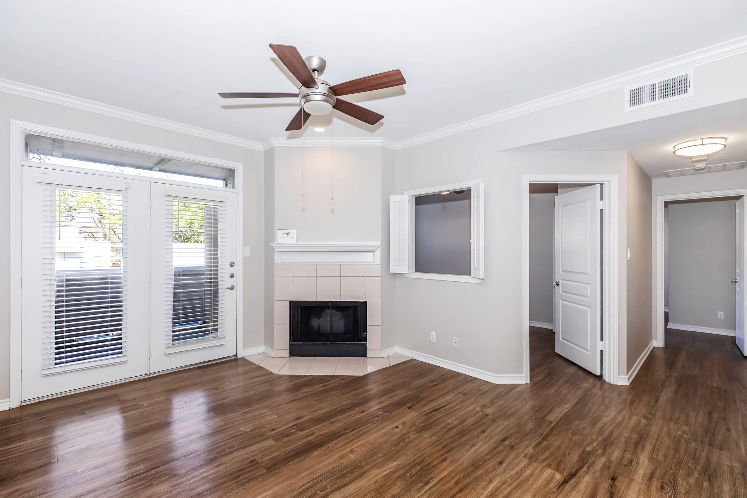 A spacious living room featuring a ceiling fan, hardwood floors, and a fireplace. There are large windows with blinds, providing natural light. The room connects to a hallway leading to additional spaces, with neutral-colored walls and modern finishes.