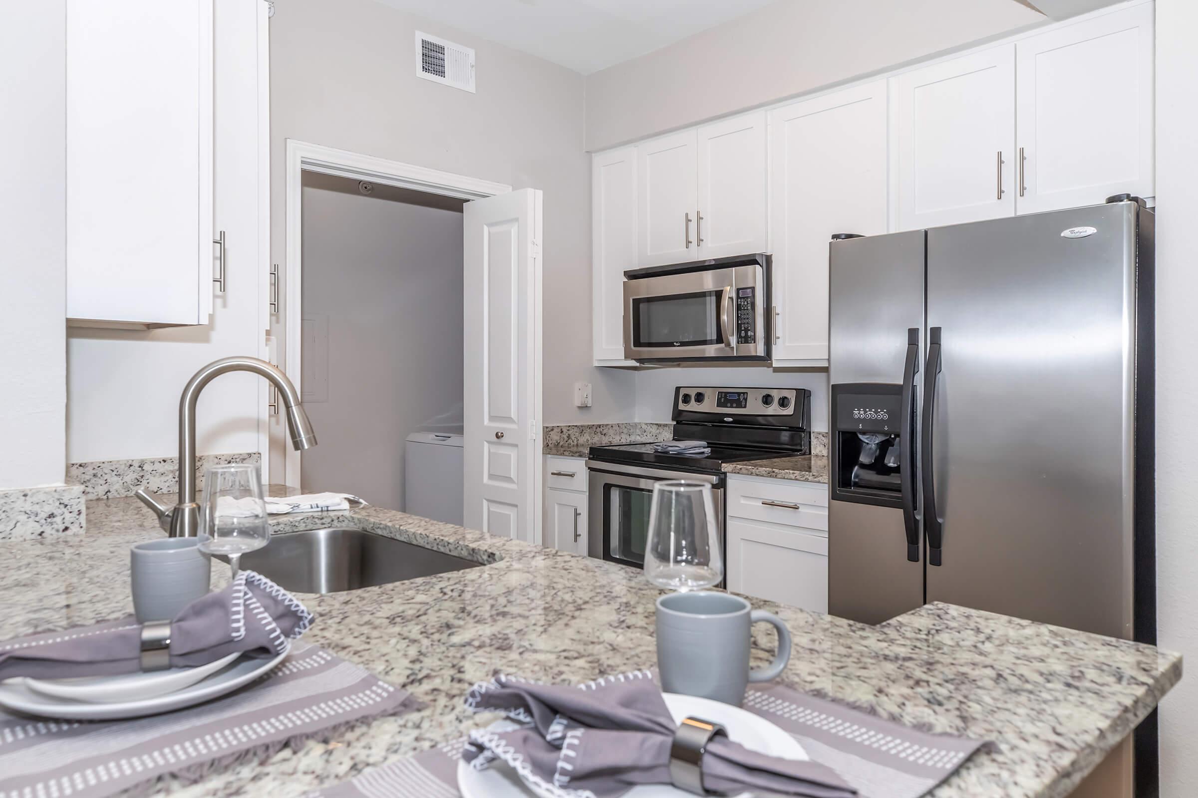 A modern kitchen featuring white cabinets, stainless steel appliances, and granite countertops. The kitchen includes a sink, microwave, stove, and refrigerator. A dining setup with gray napkins and tableware is visible, enhancing the contemporary feel of the space.