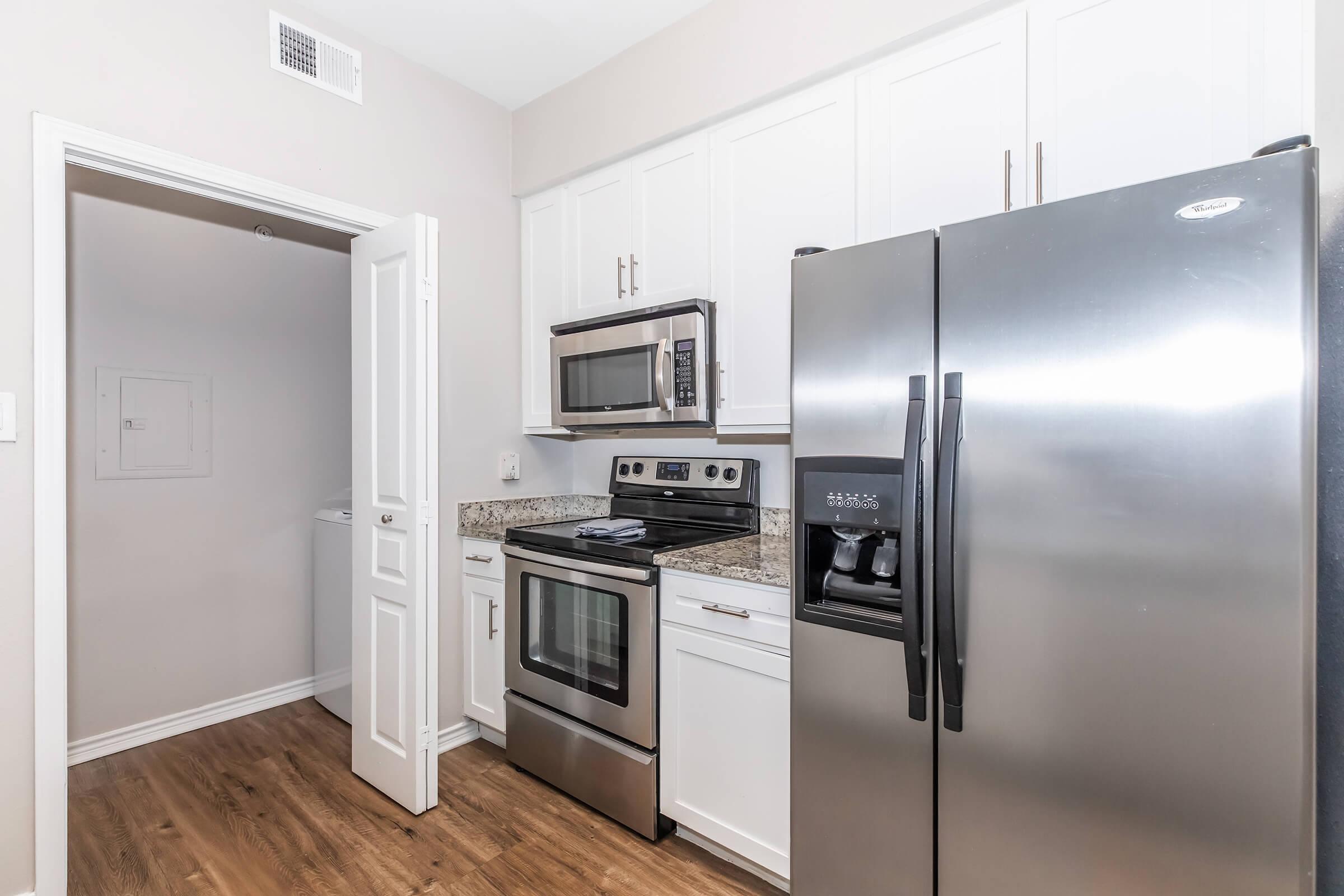 Modern kitchen featuring stainless steel appliances, including a refrigerator, microwave, and oven. Counters are topped with granite, and the cabinets are white. A doorway leads to a laundry area, and the floor has a wood-like finish. Natural light illuminates the space.