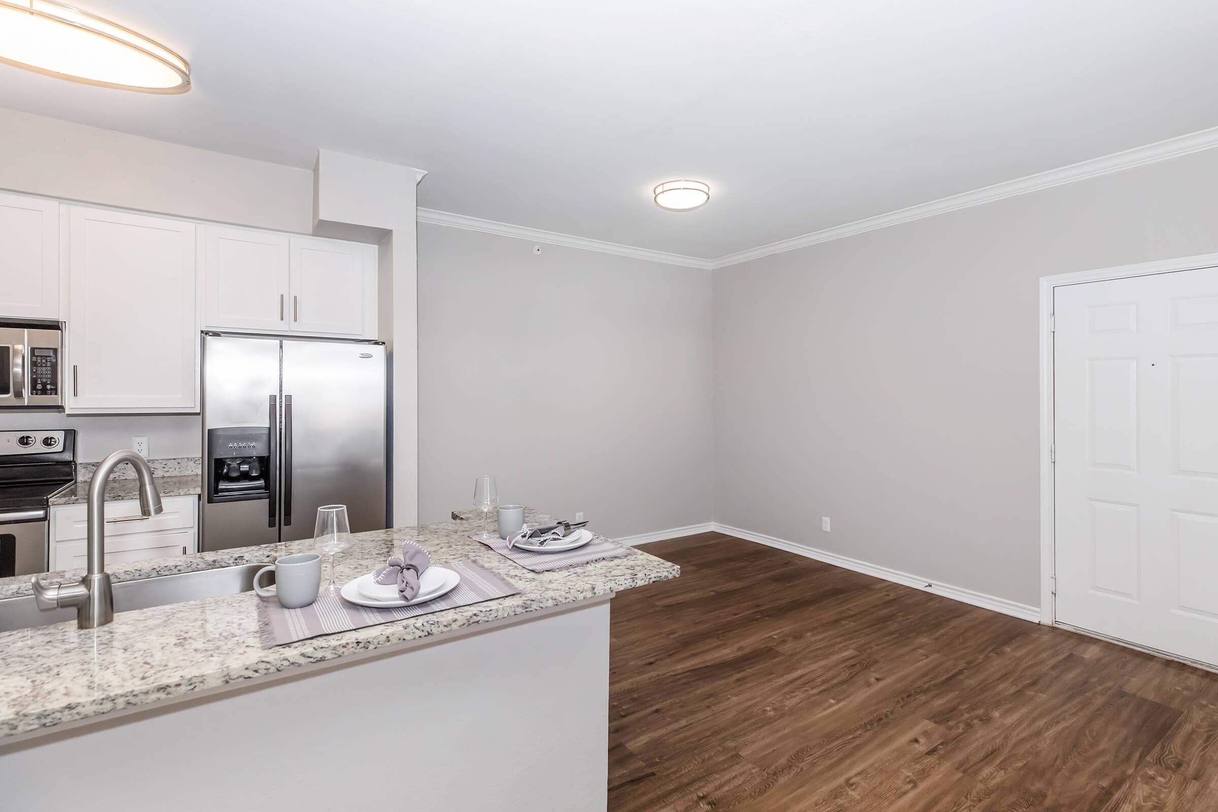 Modern kitchen and dining area featuring a granite countertop, stainless steel appliances, and a light-colored backsplash. The open space includes a dining setup with plates and glasses, hardwood flooring, and neutral-colored walls, complemented by a bright ceiling light.