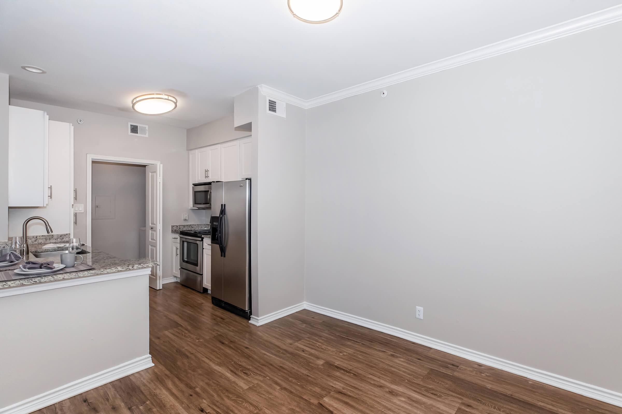 Modern kitchen with stainless steel appliances including a refrigerator and stove. The space features white cabinetry, granite countertops, and a light-colored wall. There is a dining area with wooden flooring and a door leading to another room. Brightly lit with circular ceiling fixtures.