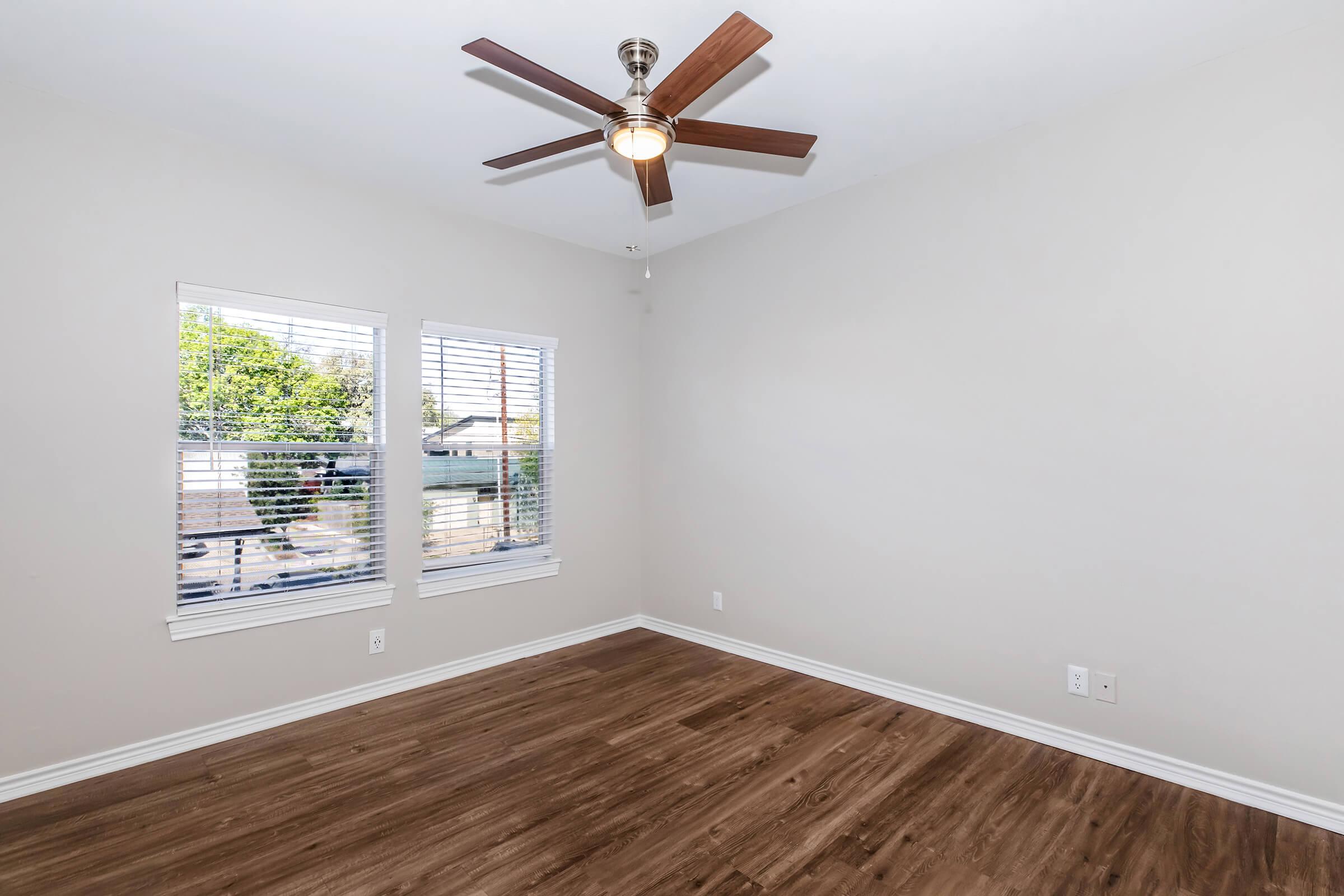 A spacious, empty room with light-colored walls and a ceiling fan. Two windows with white trim let in natural light, offering a view of greenery outside. The floor is wooden, creating a warm and inviting atmosphere. No furniture is present, highlighting the room's potential for personalization.