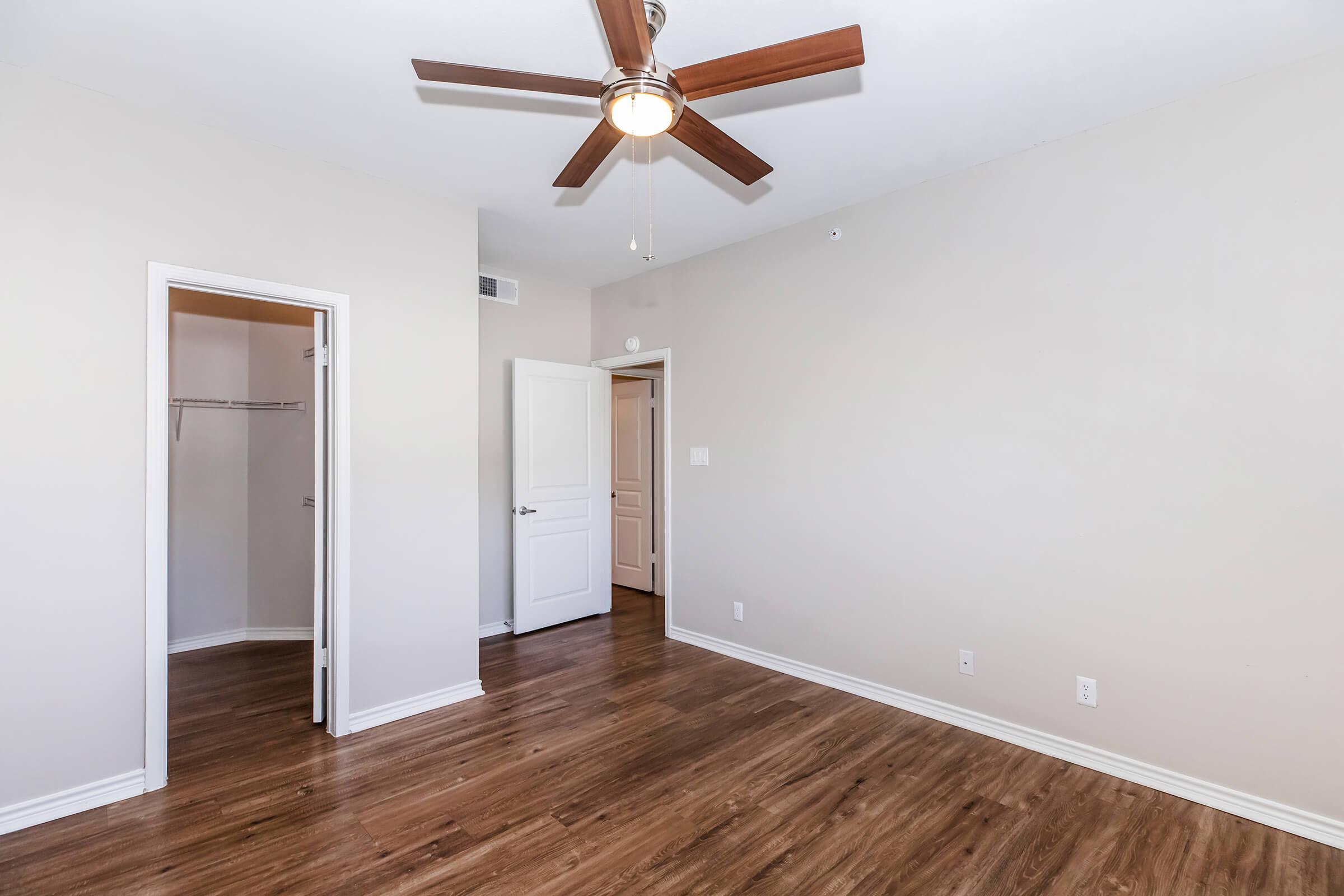 A well-lit room featuring a ceiling fan and hardwood flooring. There are two doorways, one leading to a closet and the other to an adjacent space. The walls are painted in a neutral tone, creating a spacious and inviting atmosphere.