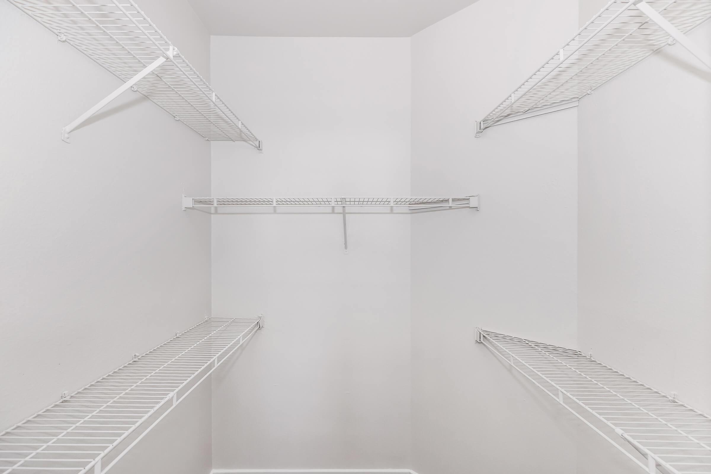 Empty closet with white wire shelving on three walls, featuring a single hanging rod in the center. The walls are painted a light color, contributing to a clean and spacious look. The floor is clear, emphasizing the organized storage potential of the shelving.