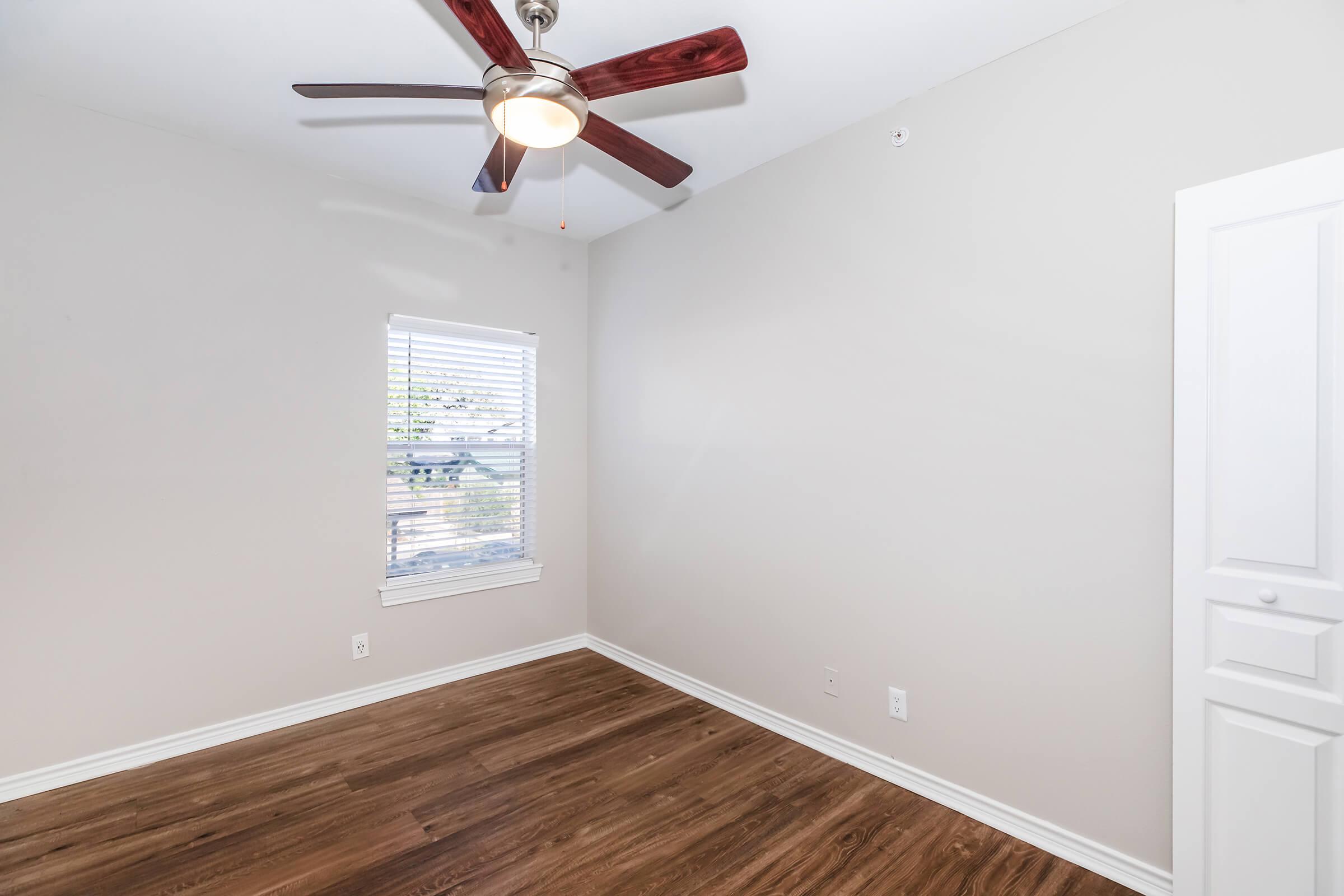 Interior view of a neutral-colored bedroom featuring a ceiling fan, a window with blinds allowing natural light, and hardwood flooring. A white closet door is visible in the corner, adding storage space. The walls are painted light gray, creating a calm and inviting atmosphere.