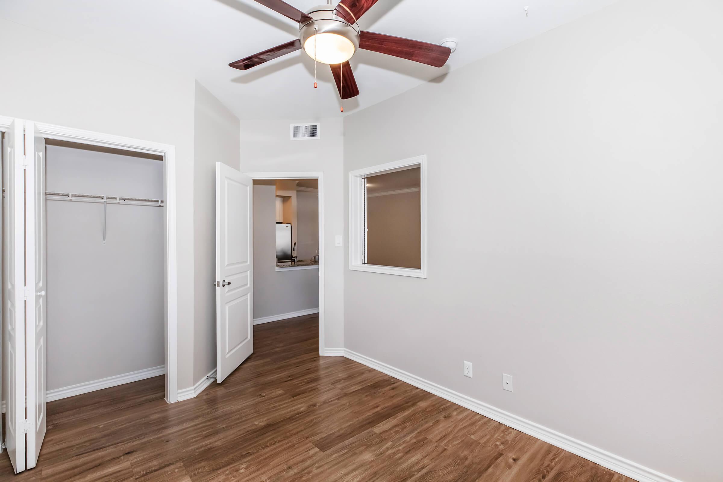 A well-lit room featuring a ceiling fan, hardwood floors, and two doorways leading to an adjoining space. One doorway opens to a closet with hanging space, while the other leads to a kitchen area visible through a window. The walls are painted in a light shade, enhancing the sense of openness.