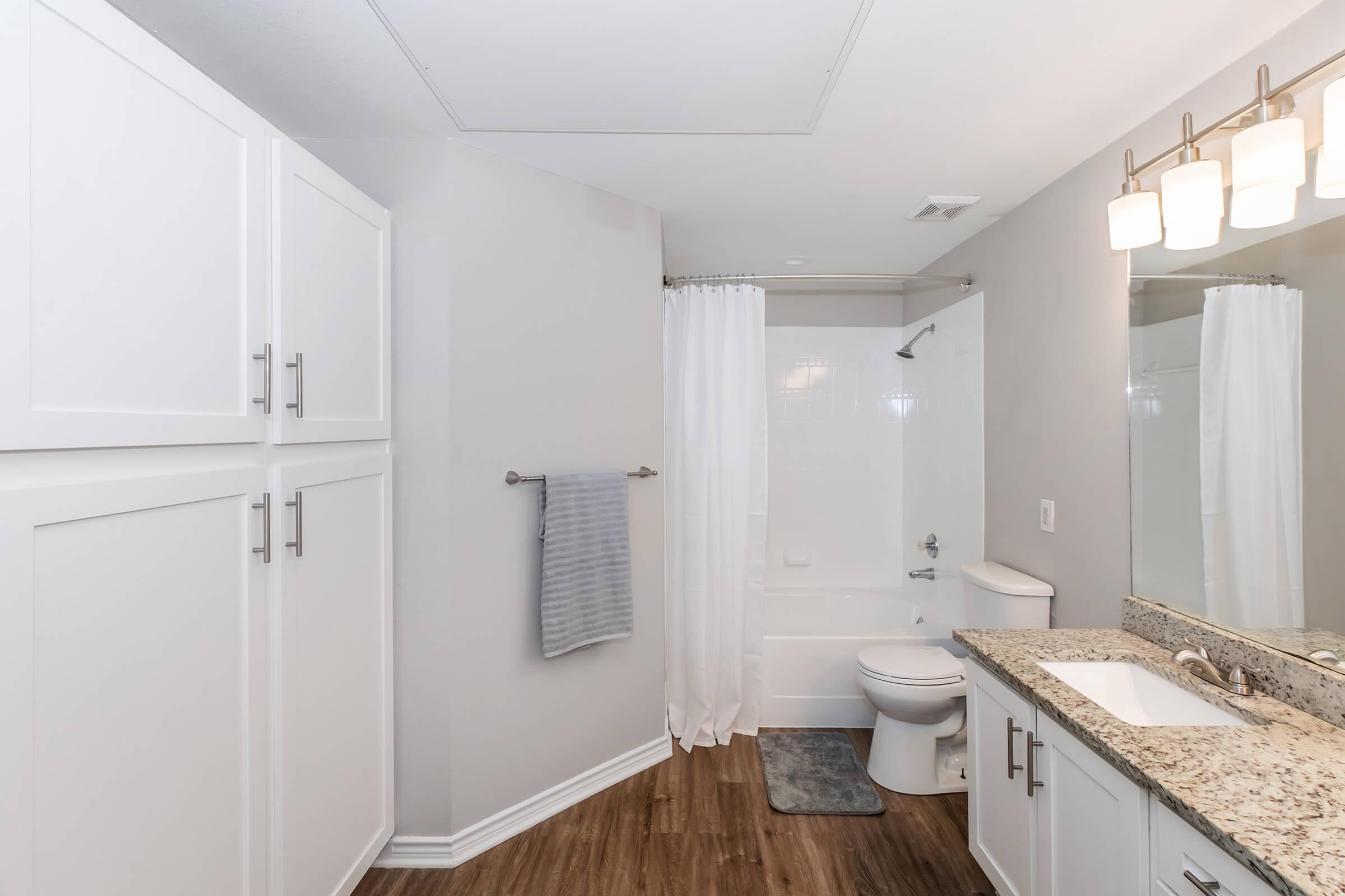 A modern bathroom featuring a granite countertop vanity with a large mirror, white cabinetry, a towel rack with a gray towel, a shower with a white curtain, and a toilet. The floor is wood-like, and the walls are painted light gray. The overall design is clean and contemporary.