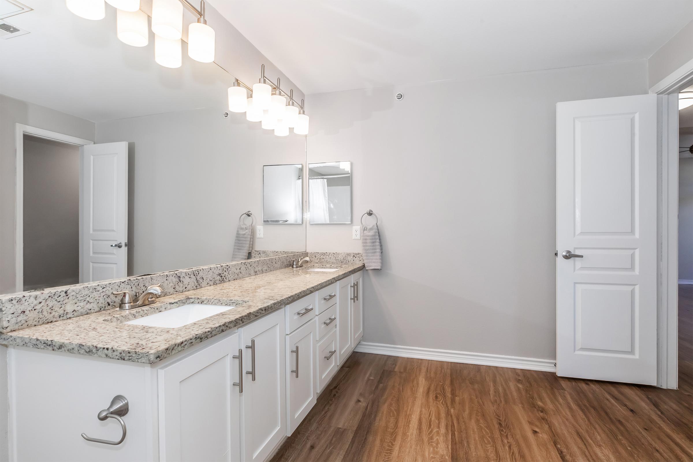 A modern bathroom featuring a long granite countertop with a white sink, under-mount faucet, and spacious storage cabinets. Light-colored walls complement the warm wood flooring. Two towels hang neatly on towel bars, while a large mirror reflects the well-lit space with contemporary light fixtures above.