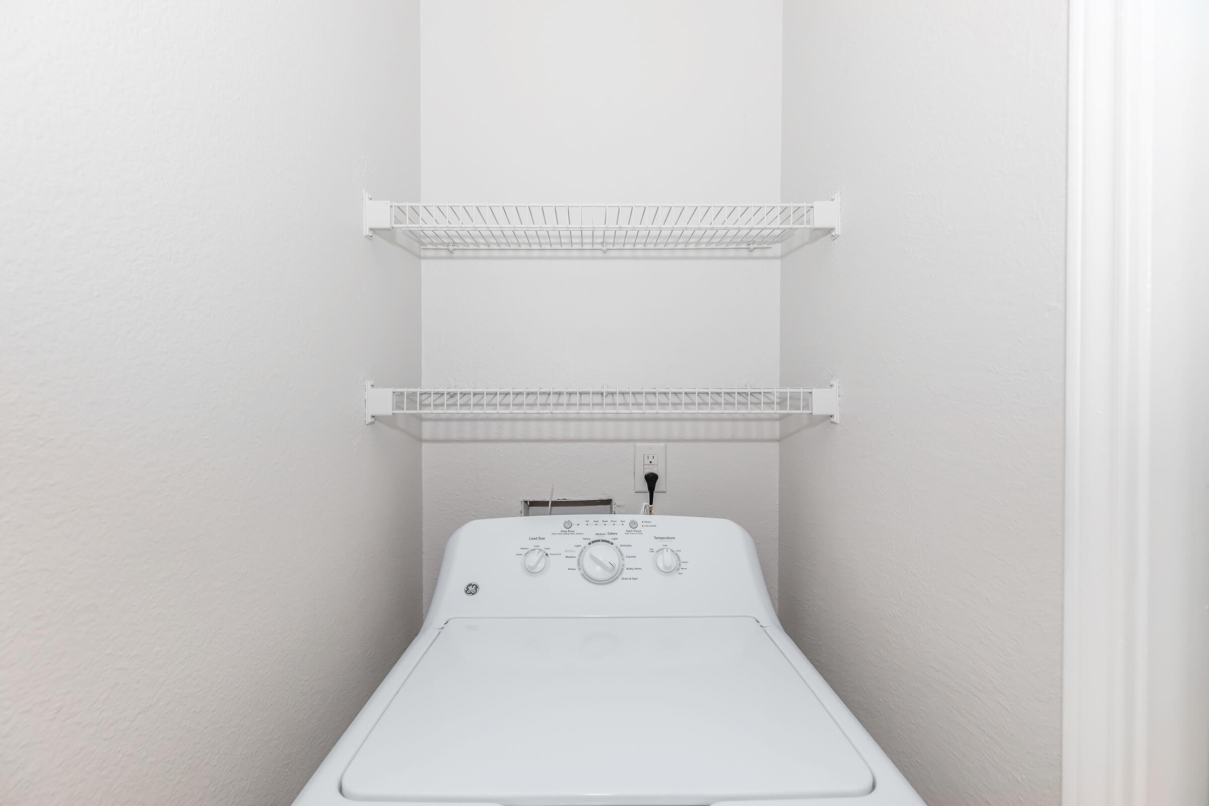 A white washing machine is situated in a small laundry space with light gray walls. Above the machine, there are two white wire shelves for additional storage. The washing machine features a control panel with knobs and buttons for various settings.