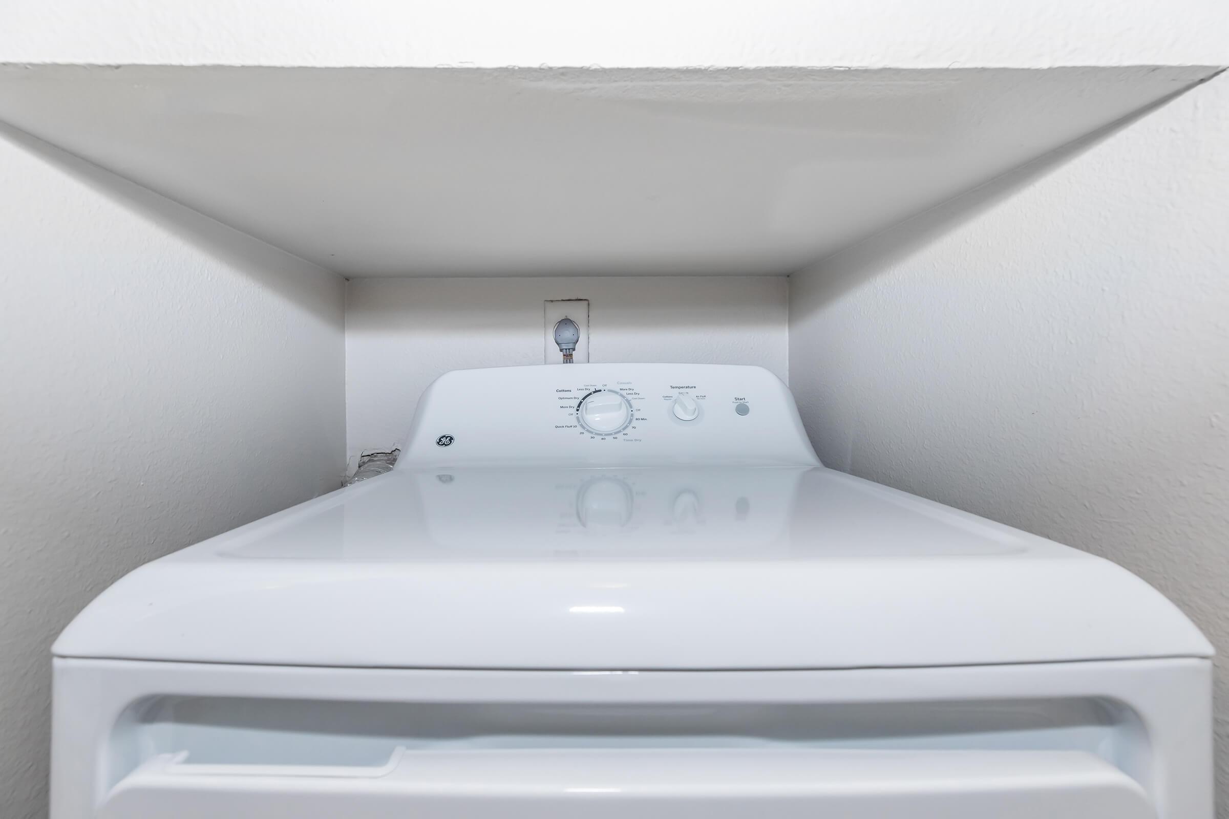 A white washing machine placed snugly in a laundry nook, with a control panel on top featuring dials and buttons for various settings. The wall above is plain and the washing machine is situated close to a power outlet.