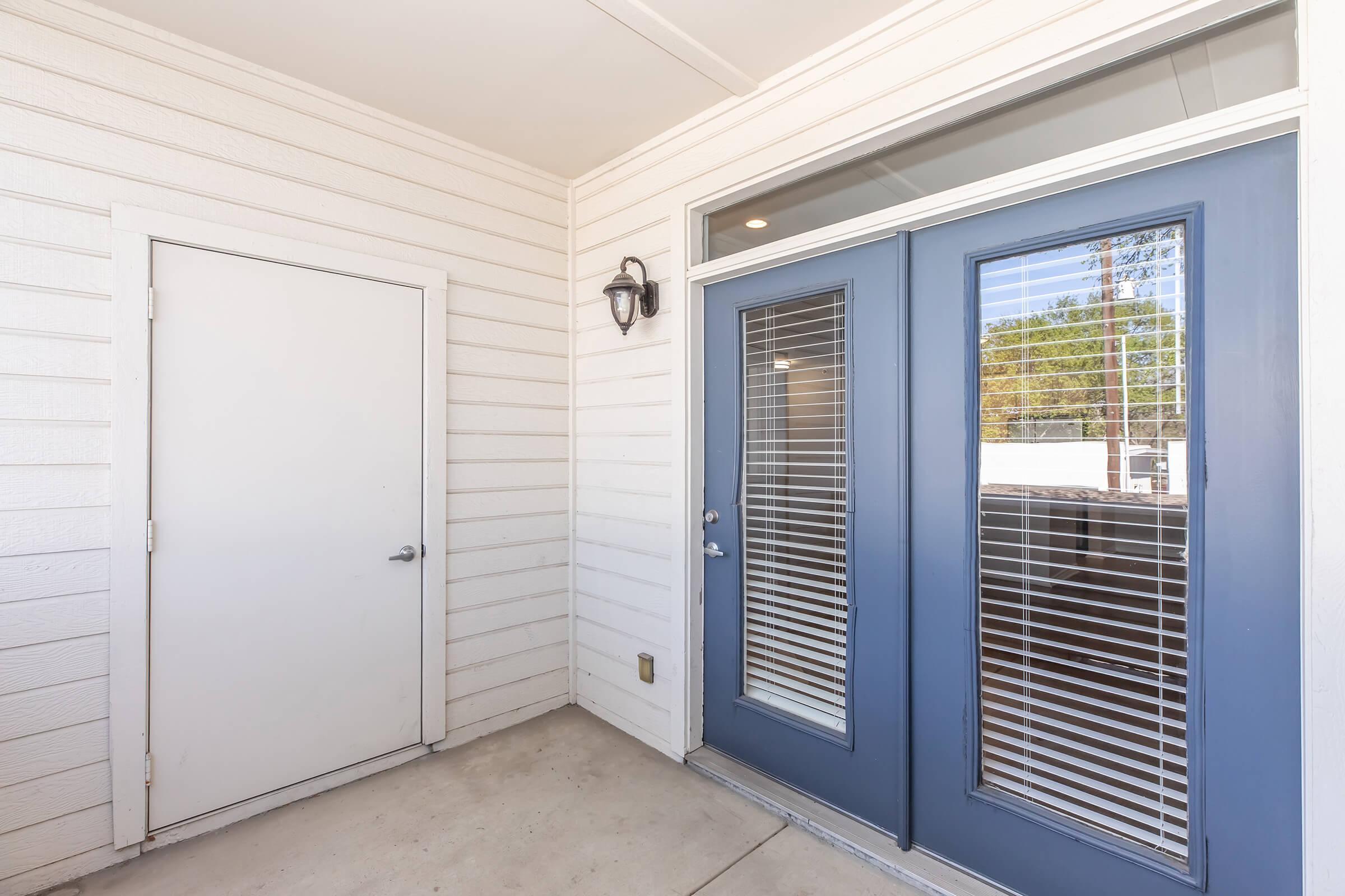A modern entryway featuring two blue double doors with blinds, and a single white door on the left. The surrounding wall is light-colored with horizontal siding, and there is a wall-mounted light fixture beside the doors. The concrete floor is clean and uncluttered.