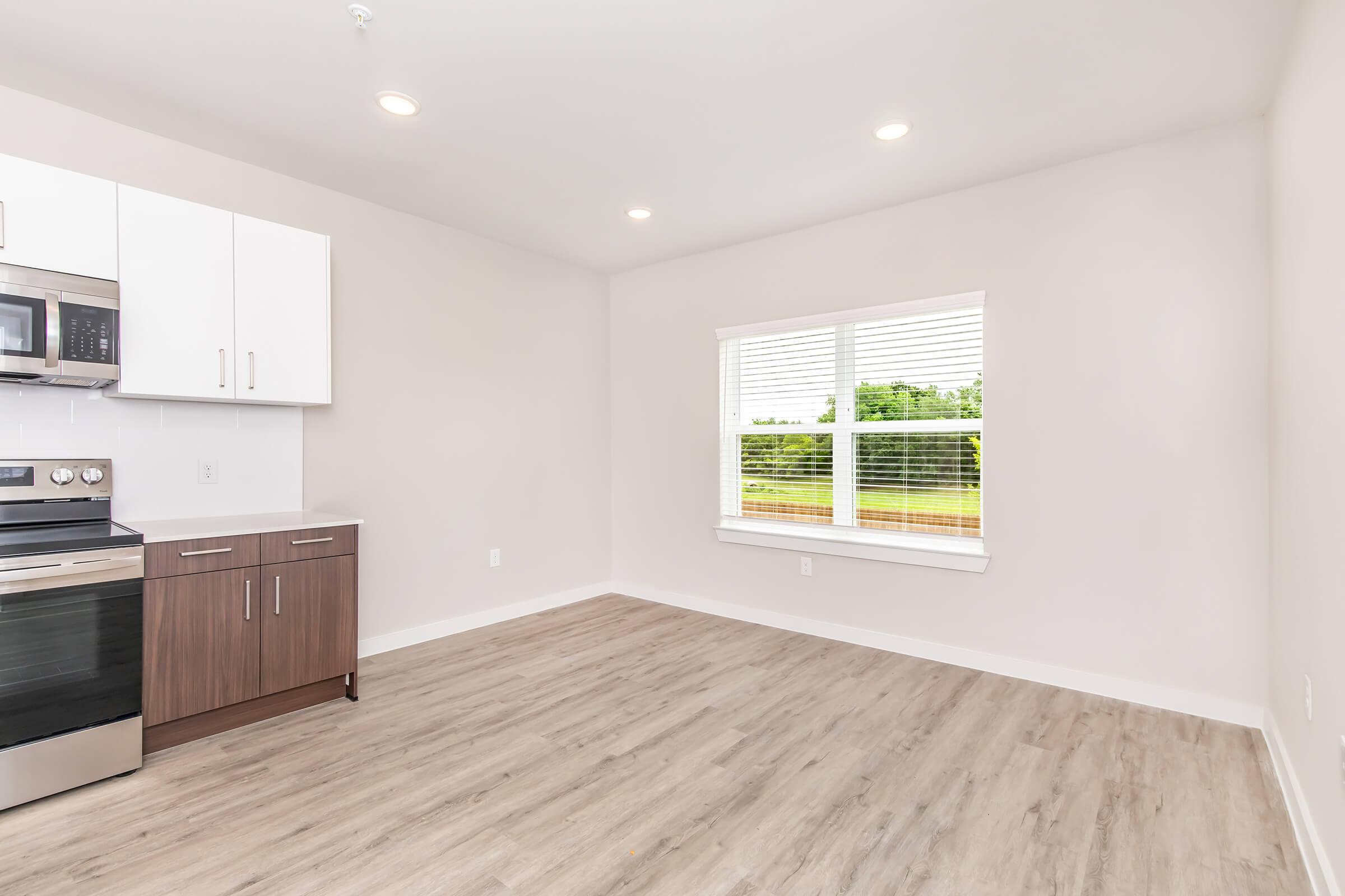 A clean, modern kitchen with stainless steel appliances, including an oven and a microwave. The kitchen features white cabinets and a wooden lower cabinet. The space is well-lit with recessed lighting and a large window that provides a view of greenery outside, complemented by light-colored walls and laminate flooring.