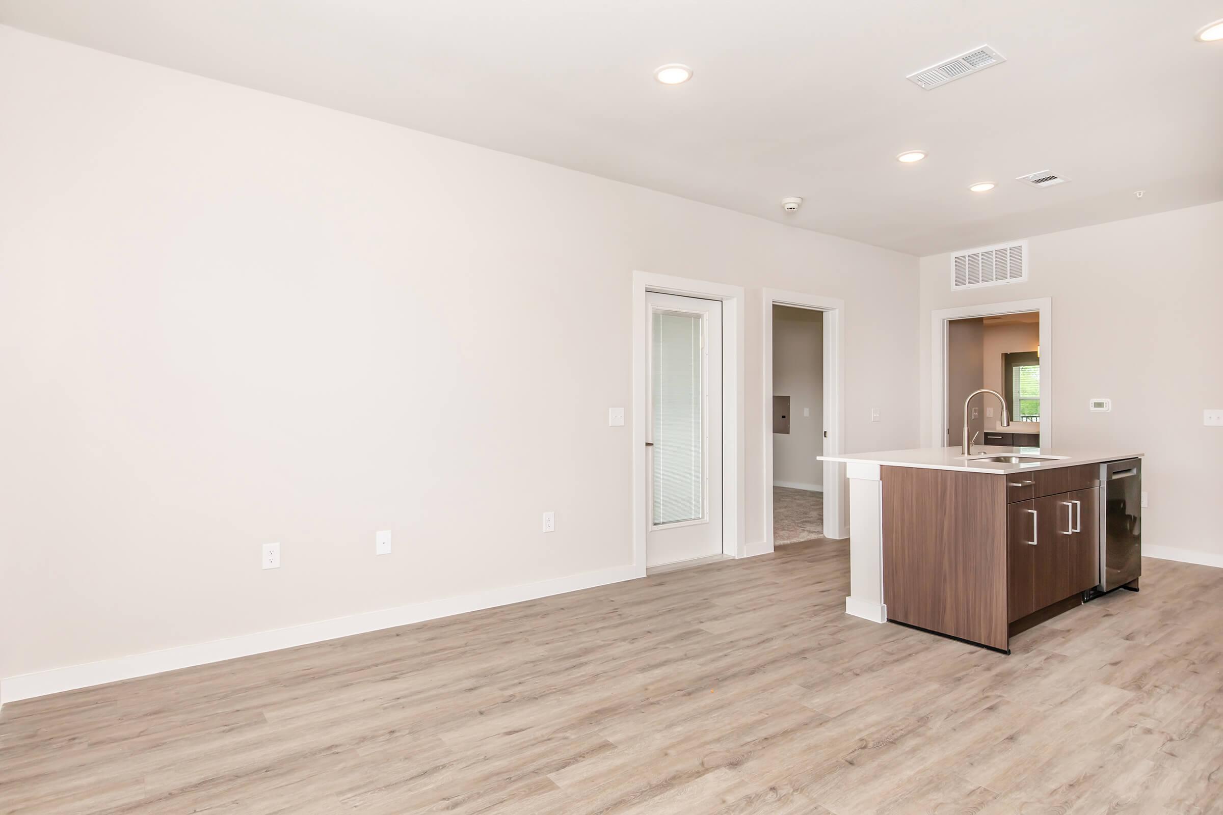 Modern, spacious living area featuring light-colored walls and wood-like flooring. A kitchen island with a sink and dark cabinetry is visible, with an open layout leading to a doorway. Natural light enters through unseen windows, creating a bright atmosphere.